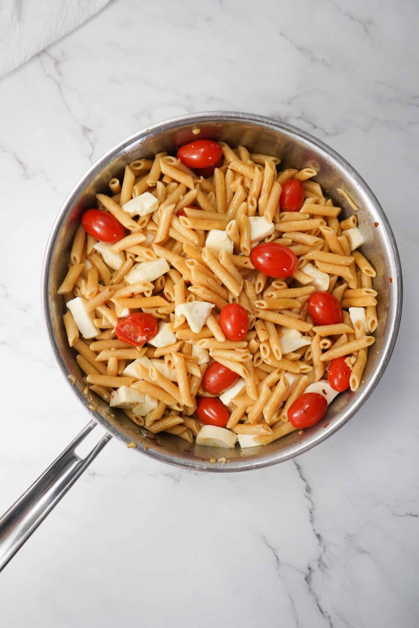 A skillet of Tuscan-inspired penne pasta, with cherry tomatoes and mozzarella chunks, sits invitingly on a marble surface.