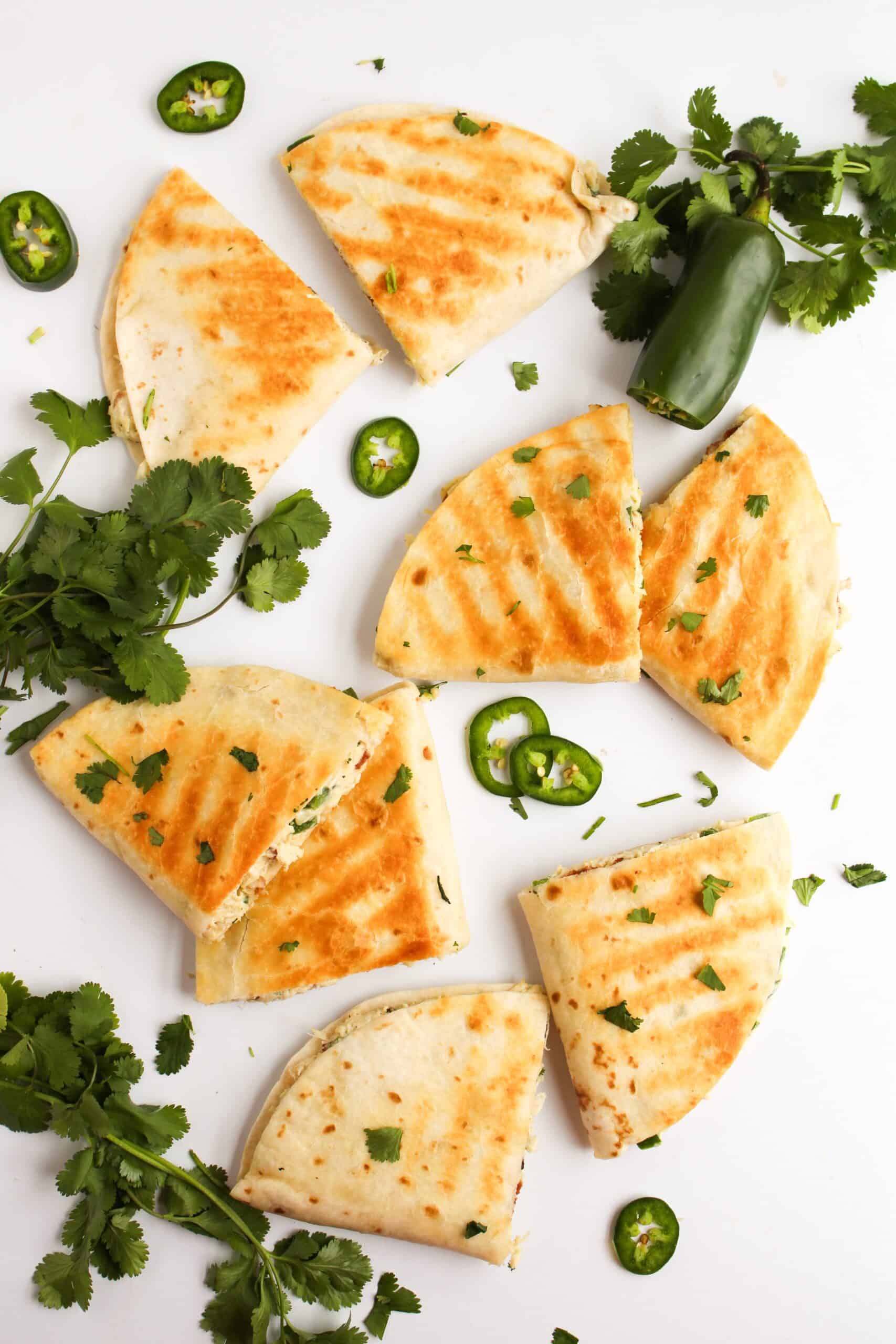 Grilled chicken quesadilla slices arranged on a white surface, surrounded by cilantro leaves and sliced jalapeños.
