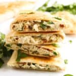 Mouthwatering stacked chicken quesadillas with a jalapeño popper twist, brimming with visible jalapeños and cilantro, surrounded by fresh cilantro leaves and sliced jalapeños on a pristine white surface.