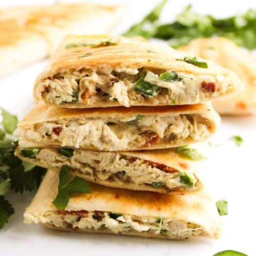 Mouthwatering stacked chicken quesadillas with a jalapeño popper twist, brimming with visible jalapeños and cilantro, surrounded by fresh cilantro leaves and sliced jalapeños on a pristine white surface.