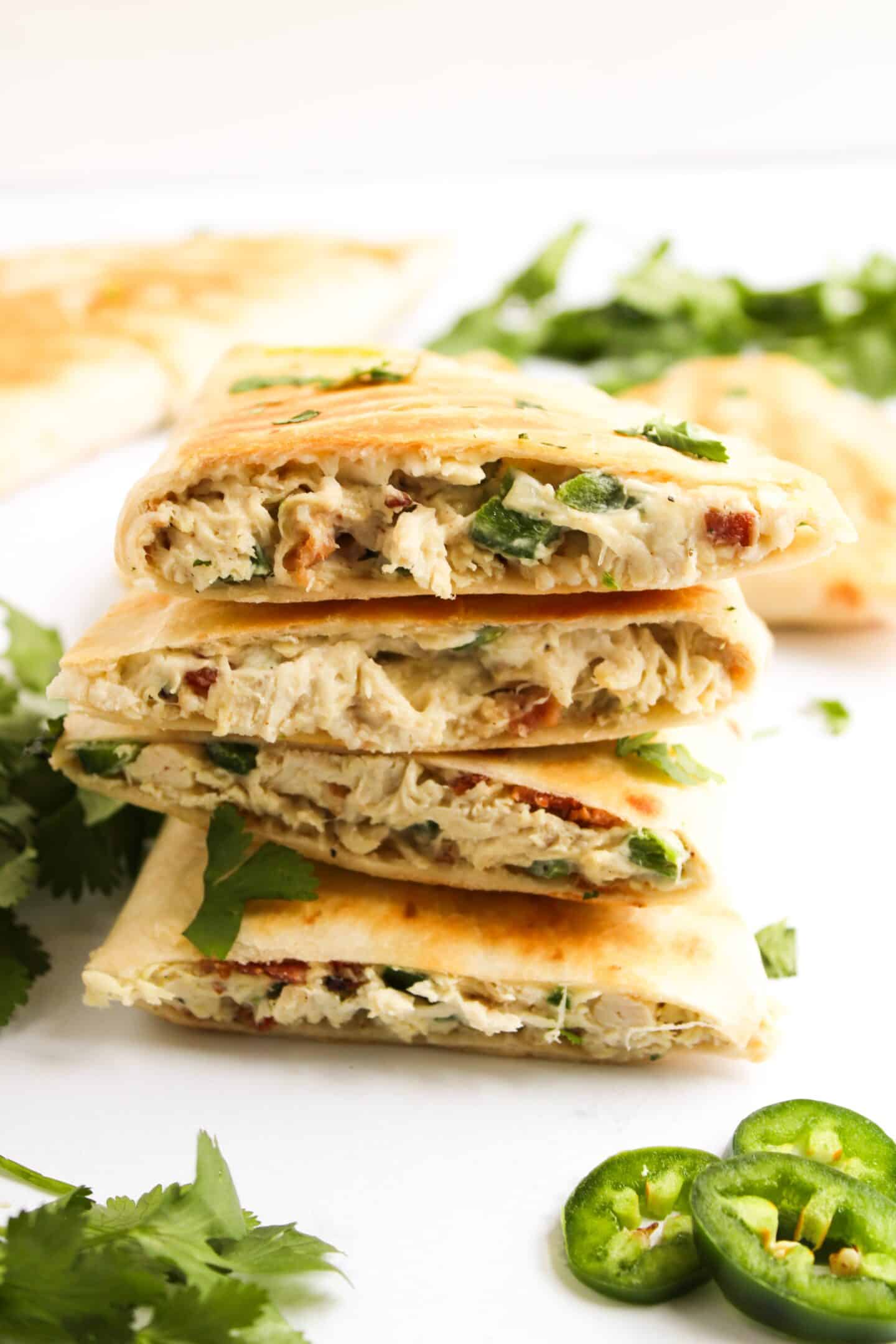 Stacked chicken quesadillas, reminiscent of a jalapeño popper, reveal inviting layers of chicken, herbs, and jalapeño slices. They rest elegantly on a white surface, encircled by fresh cilantro and even more jalapeño slices.