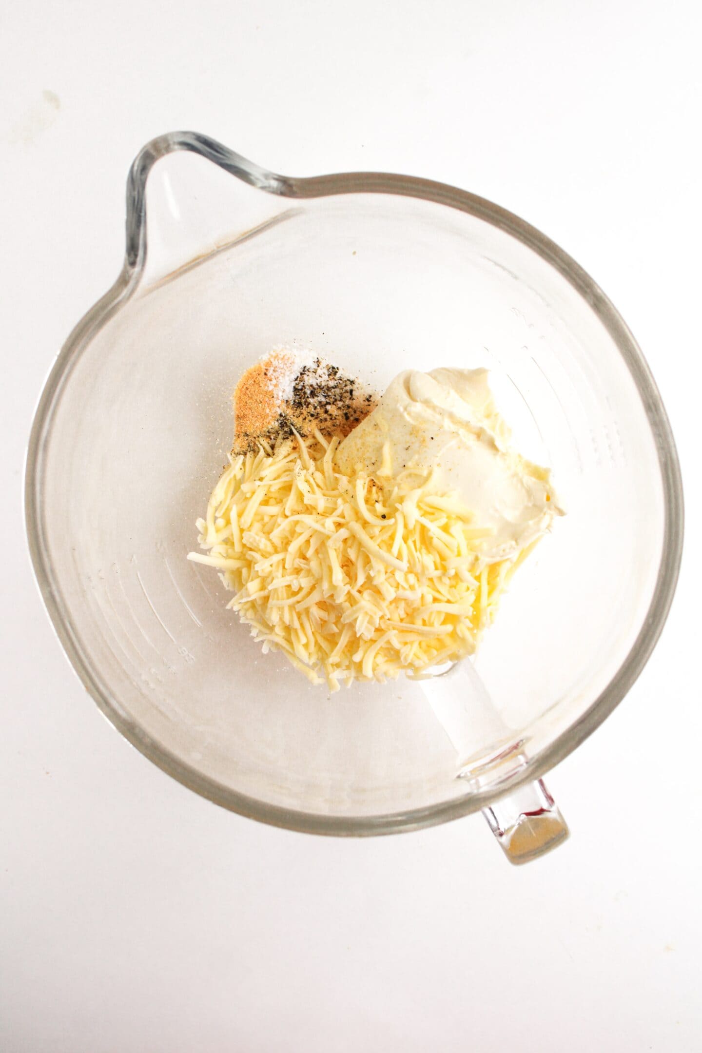 A glass bowl contains shredded cheese, mayonnaise, spices, and a dollop of mustard on a white background—perfect ingredients to spice up your jalapeño popper chicken quesadillas.