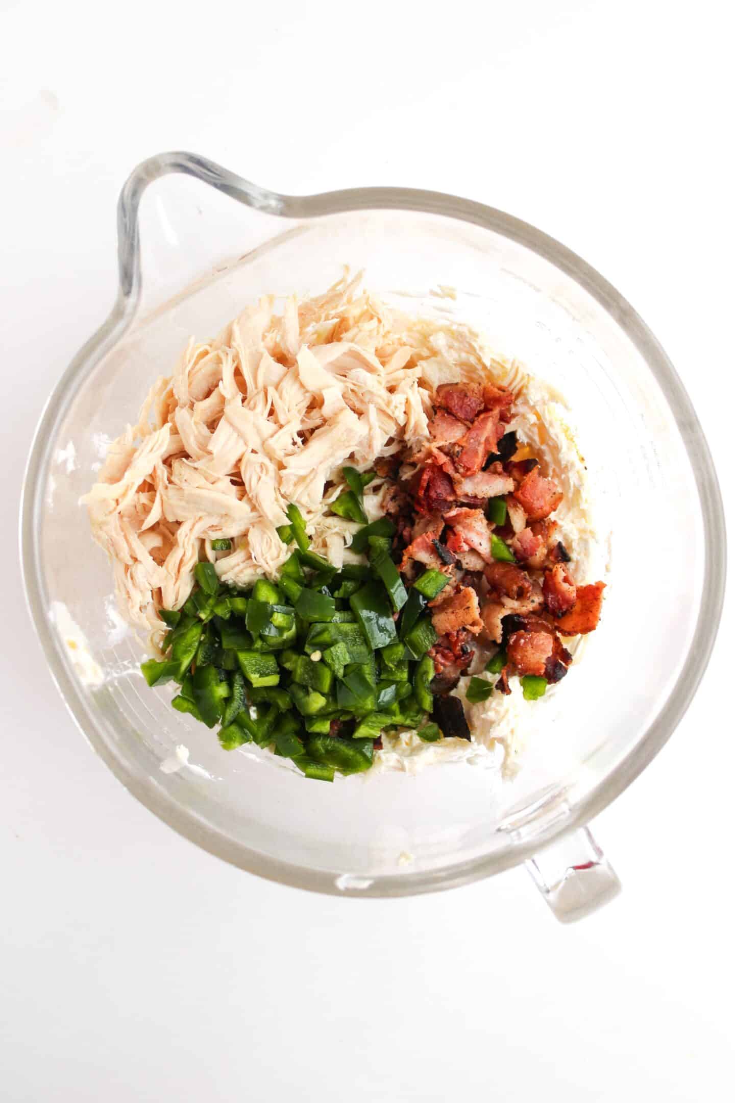 A glass bowl featuring the enticing flavors of Jalapeño Popper Chicken, with shredded chicken, chopped jalapeños, and crispy bacon pieces on a white surface.