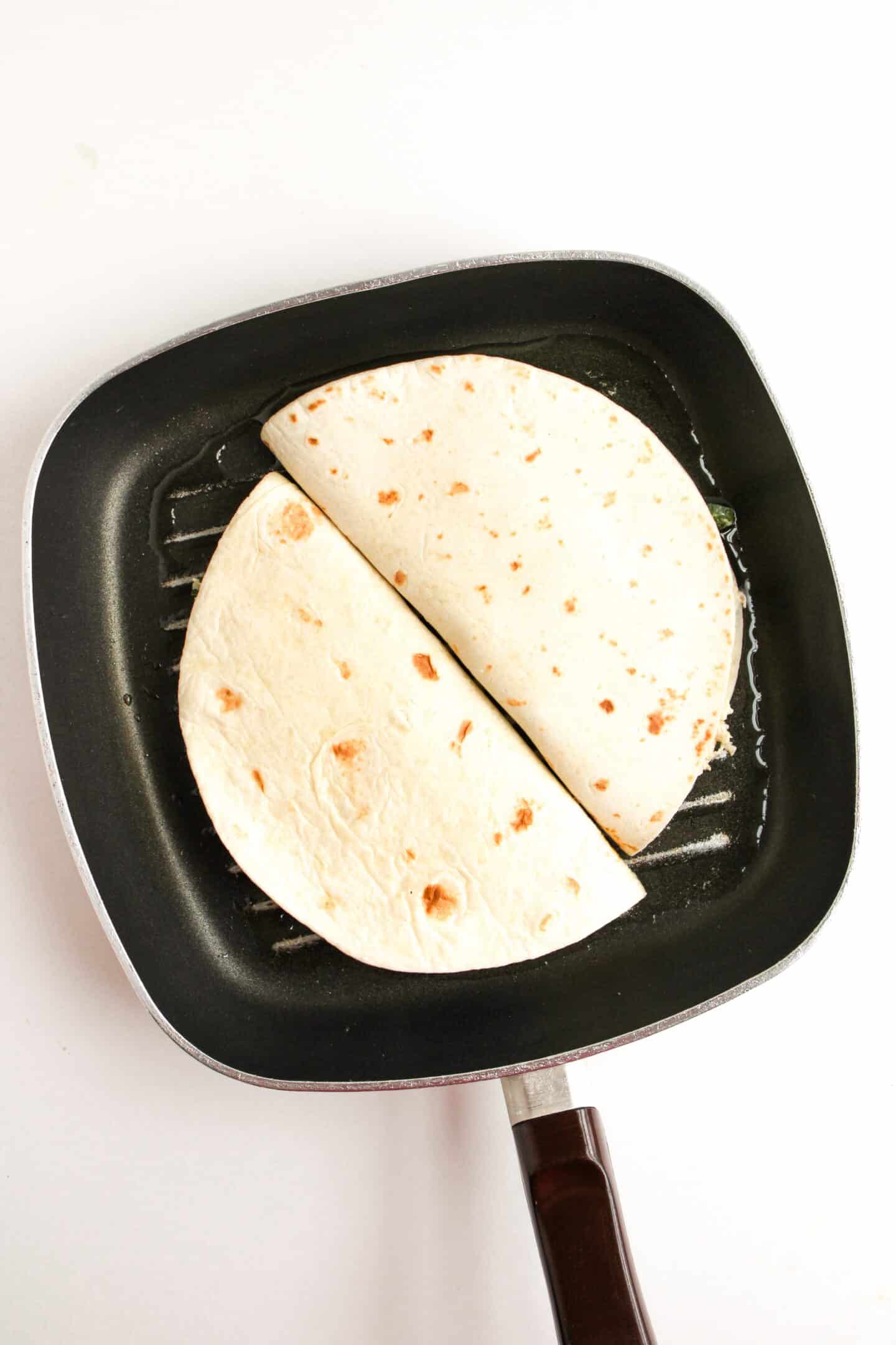 Two folded quesadillas heat on a black grill pan with a wooden handle, filling the air with their tempting aroma.