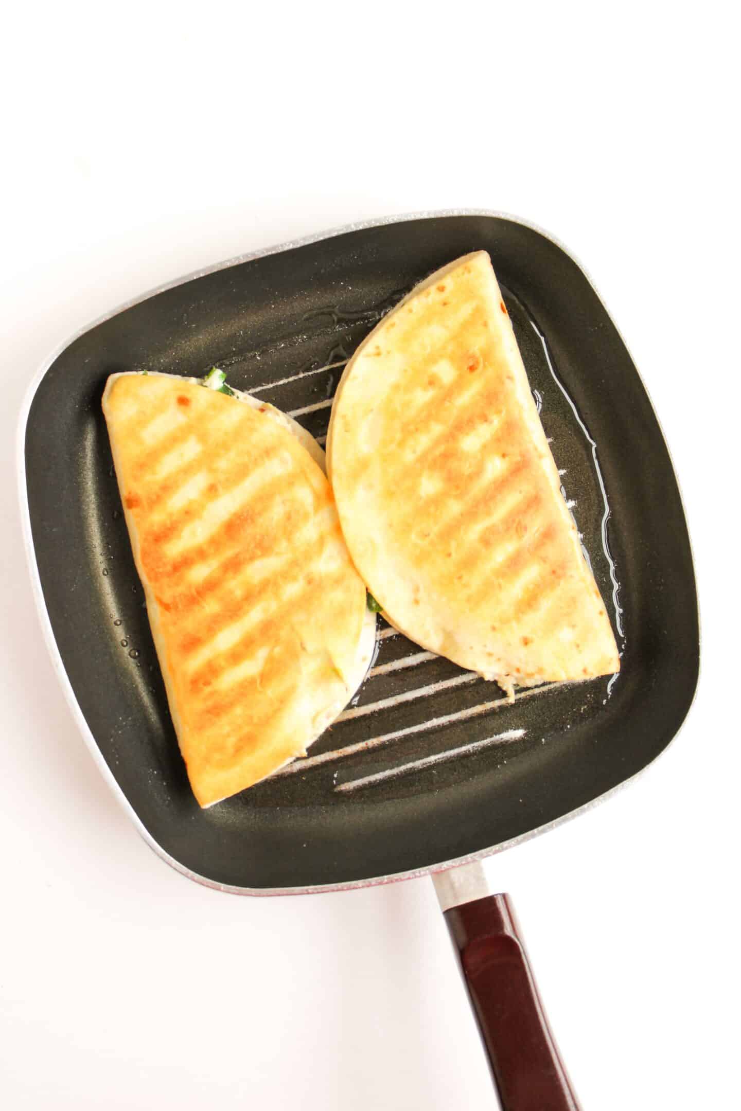 Sizzling in the frying pan on a pristine white background are two perfectly grilled Jalapeño Popper Chicken Quesadillas, tempting your taste buds with their flavorful aroma.