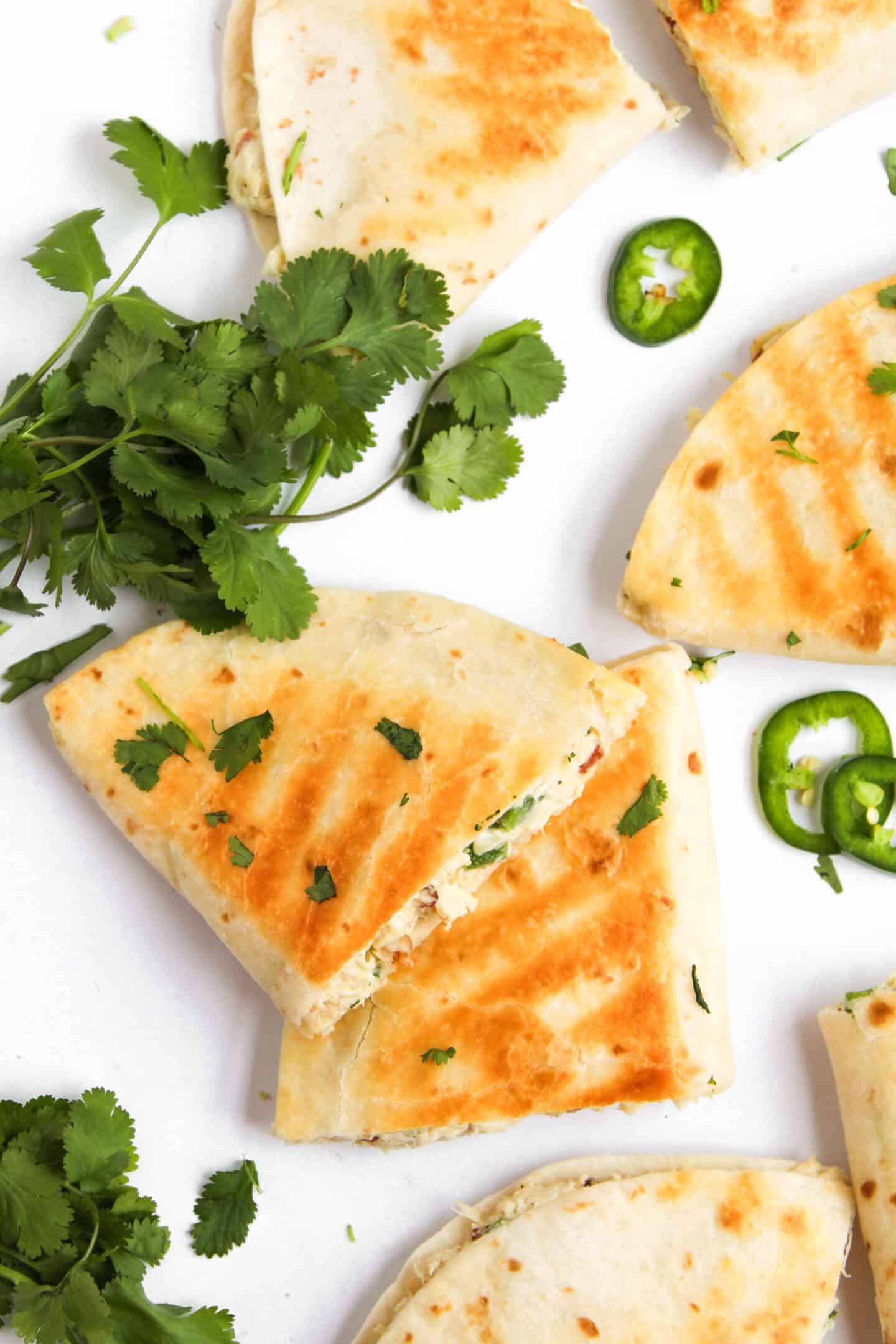 Golden brown jalapeño popper chicken quesadillas stuffed with cheese and herbs, garnished with cilantro and sliced jalapeños, on a pristine white background.