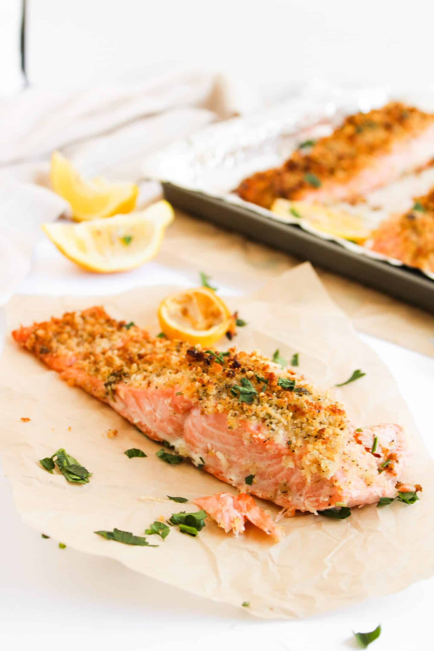 A panko-crusted baked salmon fillet, adorned with breadcrumbs and herbs, rests on parchment paper beside lemon wedges. Another salmon piece sizzles on a baking tray in the background.