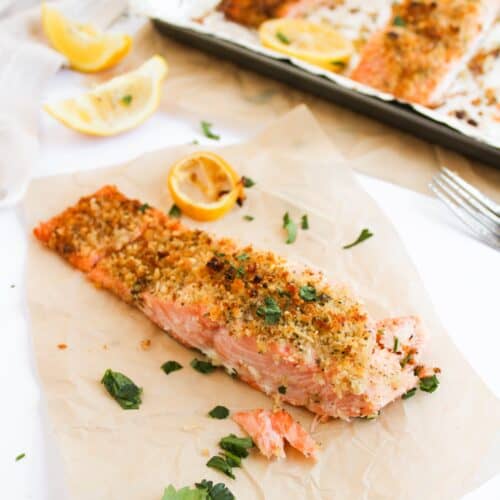 Baked salmon fillet with a panko-crusted topping of breadcrumbs and herbs rests on parchment paper. It’s adorned with lemon wedges and parsley, sitting next to a tray of more savory baked salmon pieces, ideal for any delicious salmon recipe.