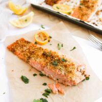 Baked salmon fillet with a panko-crusted topping of breadcrumbs and herbs rests on parchment paper. It’s adorned with lemon wedges and parsley, sitting next to a tray of more savory baked salmon pieces, ideal for any delicious salmon recipe.