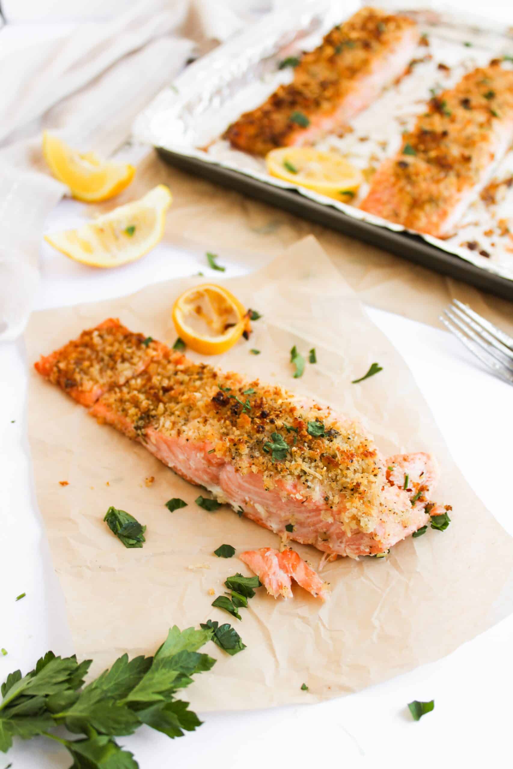 Baked salmon fillet with a panko-crusted topping of breadcrumbs and herbs rests on parchment paper. It’s adorned with lemon wedges and parsley, sitting next to a tray of more savory baked salmon pieces, ideal for any delicious salmon recipe.