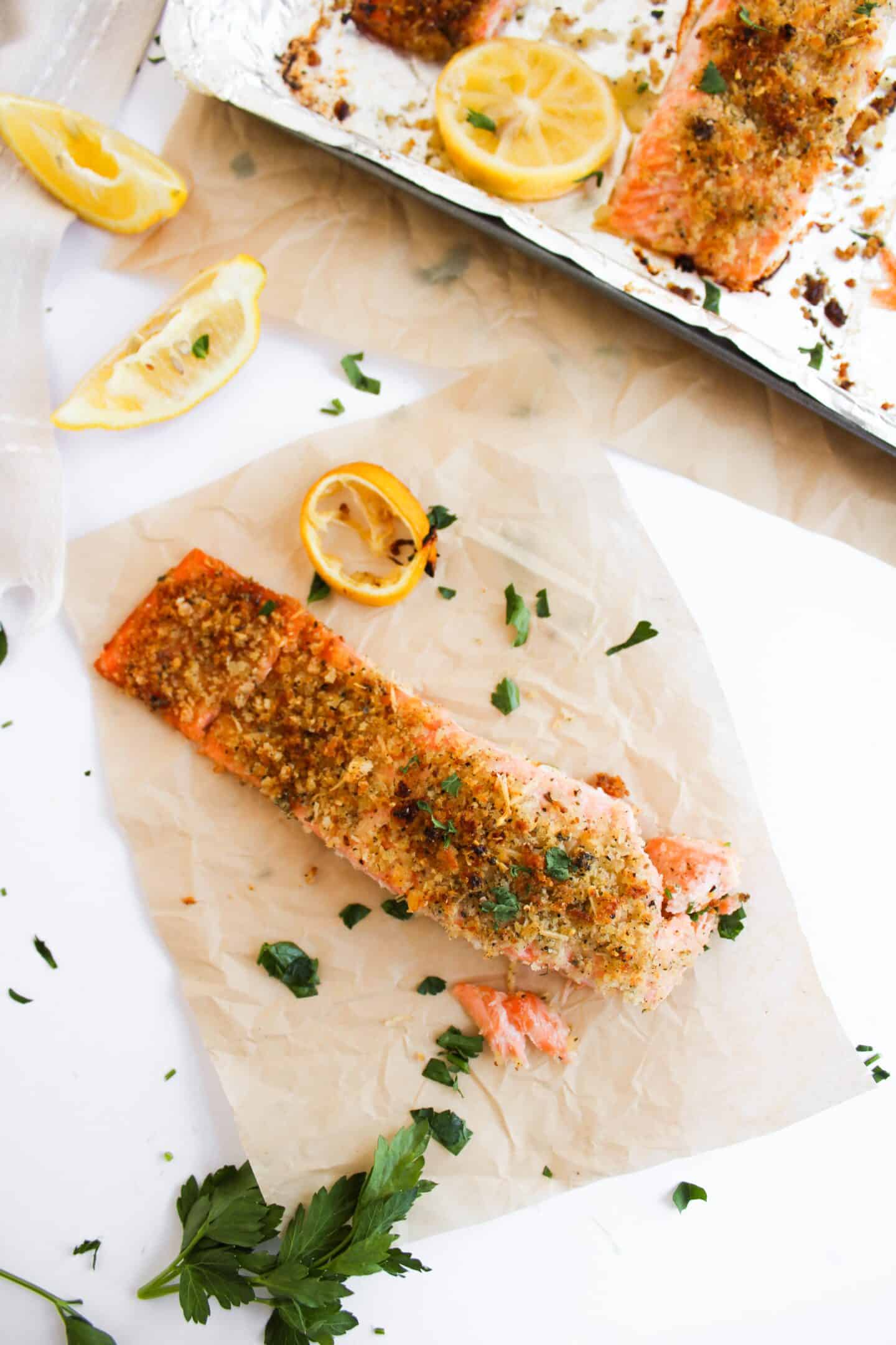 Panko-crusted baked salmon fillet sits elegantly on parchment paper, garnished with lemon wedges and parsley. In the background, another piece of salmon peeks out from a baking tray, inviting you to savor this delectable salmon recipe.
