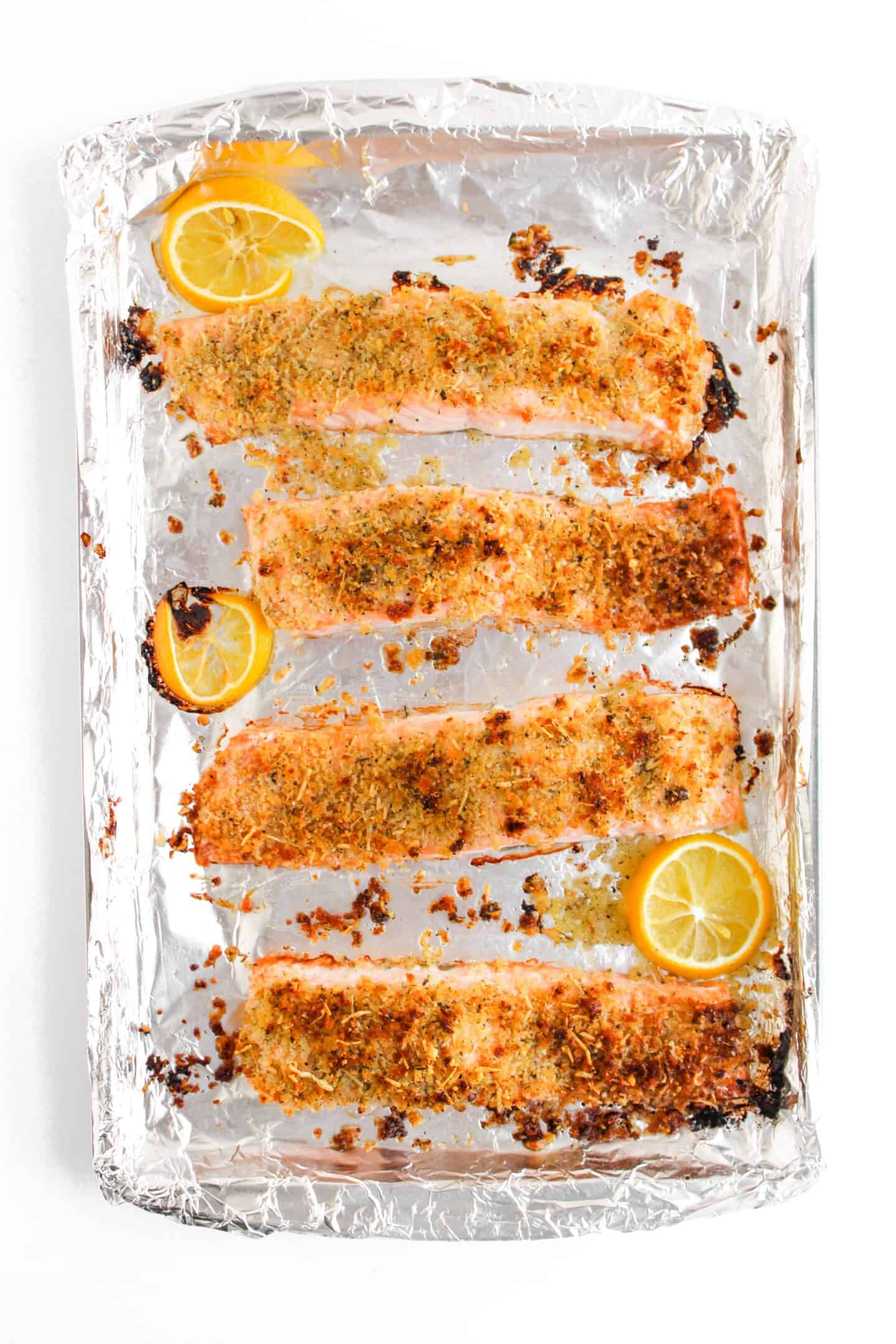 Baked salmon fillets with a panko-crusted herb topping rest on a foil-lined sheet, elegantly garnished with fresh lemon slices.