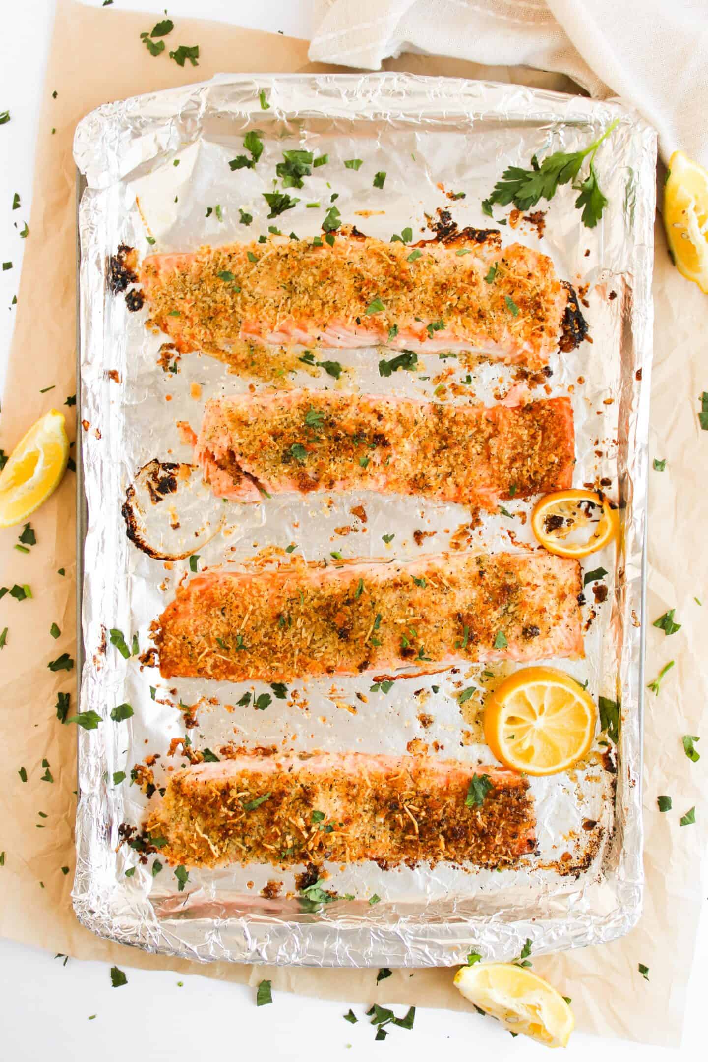 Four panko-crusted salmon fillets are garnished with parsley and lemon slices, then placed perfectly on a foil-lined baking sheet, ready for a delicious bake.