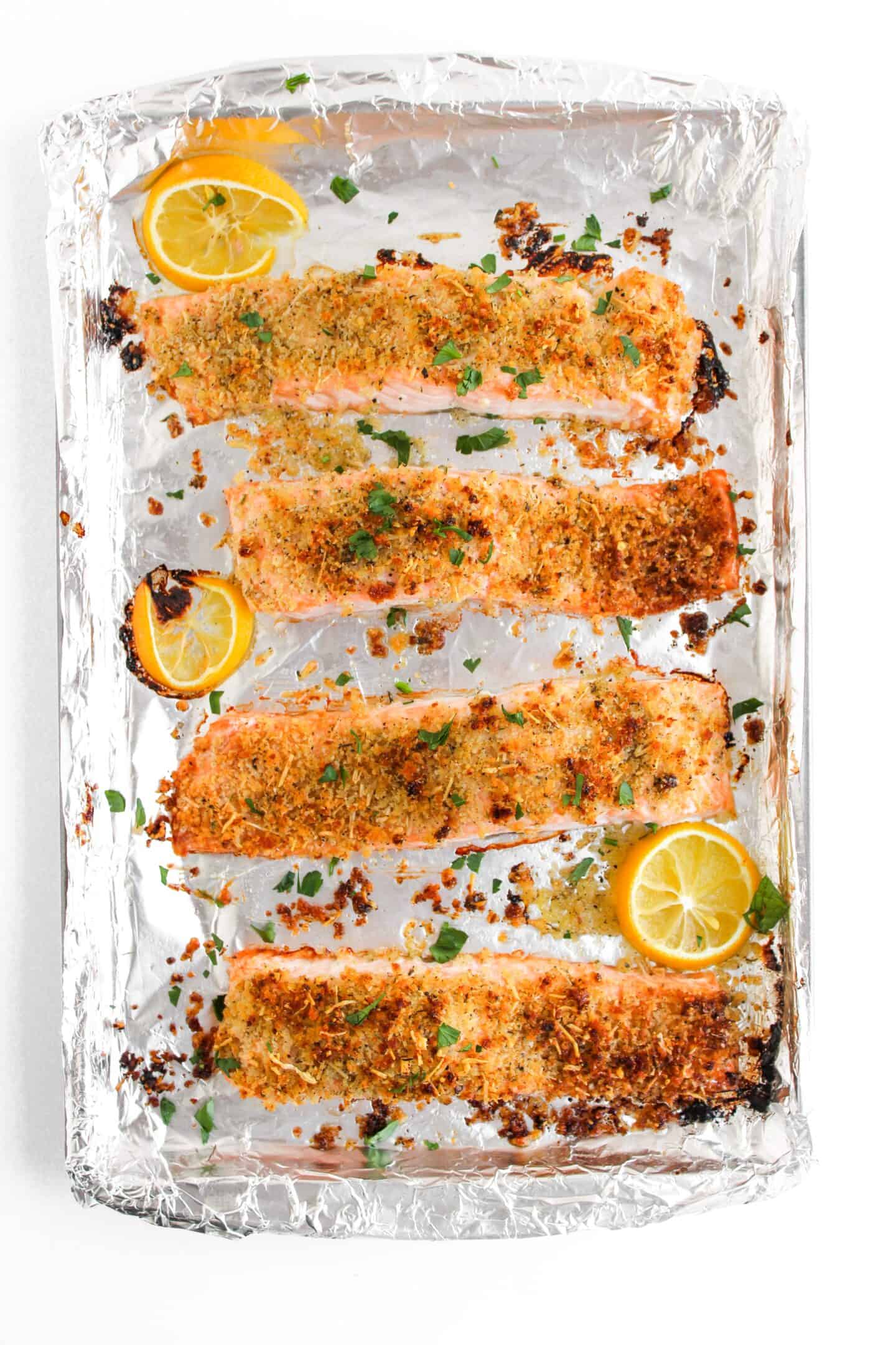 Four panko-crusted salmon fillets rest on a foil-lined baking tray, garnished with lemon slices and fresh herbs—a simple yet exquisite baked salmon recipe.