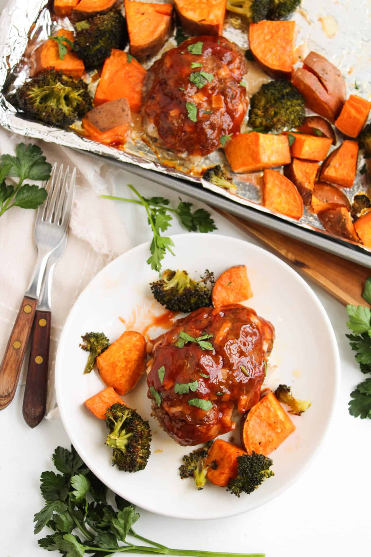 A plate with barbecue-glazed mini meatloaf, roasted sweet potatoes, and broccoli next to a fork and knife. The same items are perfectly arranged on a sheet pan in the background. Parsley garnishes the dish.