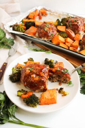 Plate with mini meatloaf glazed in sauce, surrounded by roasted veggies like broccoli and sweet potatoes. A similar sheet pan dish is visible on a nearby baking tray.