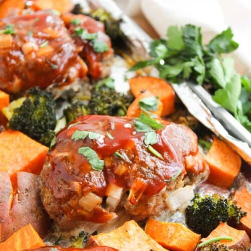 The sheet pan boasts mini meatloaf slices, each glazed with sauce, nestled among roasted sweet potatoes and broccoli. Garnished with fresh parsley sprigs, this colorful meal is brimming with vibrant veggies.