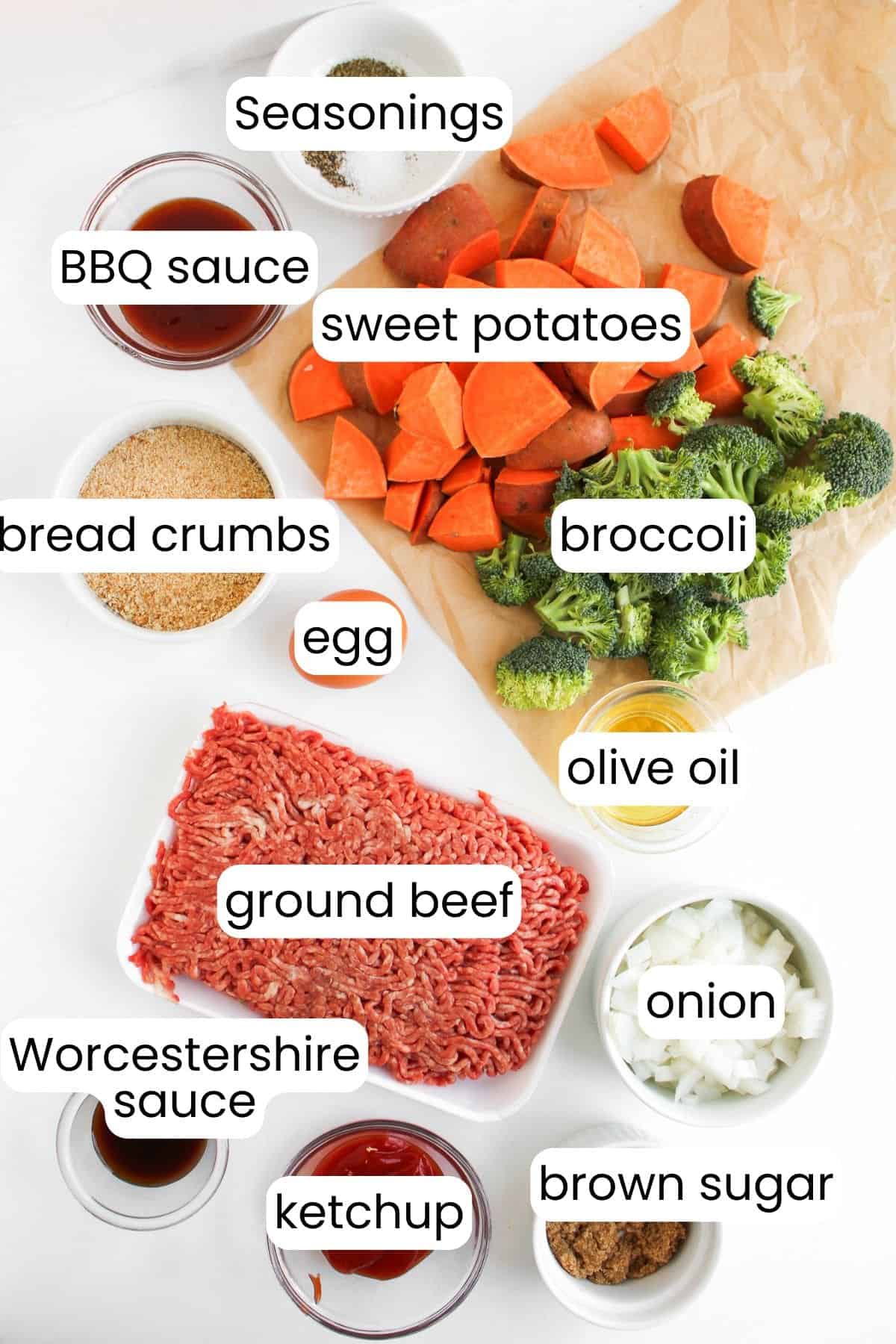 Ingredients for mini meatloaf including ground beef, sweet potatoes, and broccoli are laid out on a white surface. BBQ sauce, seasonings, bread crumbs, eggs, olive oil, onion, Worcestershire sauce, ketchup, and brown sugar ready to create a delightful sheet pan meal with veggies.