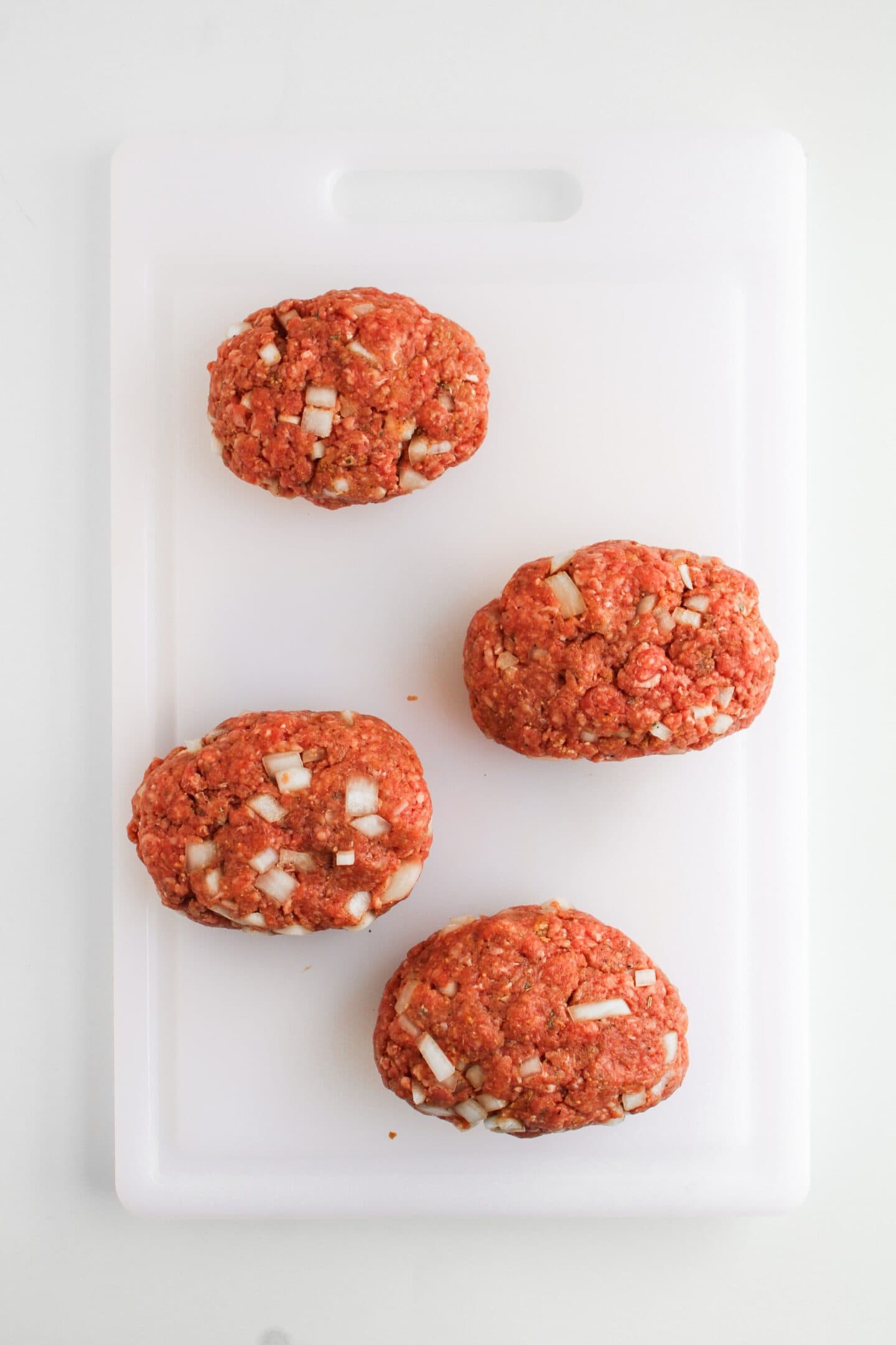 Four mini meatloaves made from raw beef with visible onion pieces are artfully placed on a white cutting board, ready to be accompanied by a colorful array of veggies for a perfect sheet pan dinner.