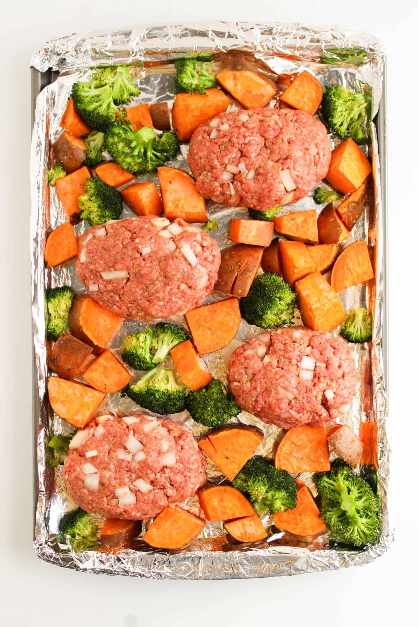 Mini meatloaf patties with diced onions, surrounded by vibrant veggies like chopped sweet potatoes and broccoli, are arranged on a sheet pan lined with foil.