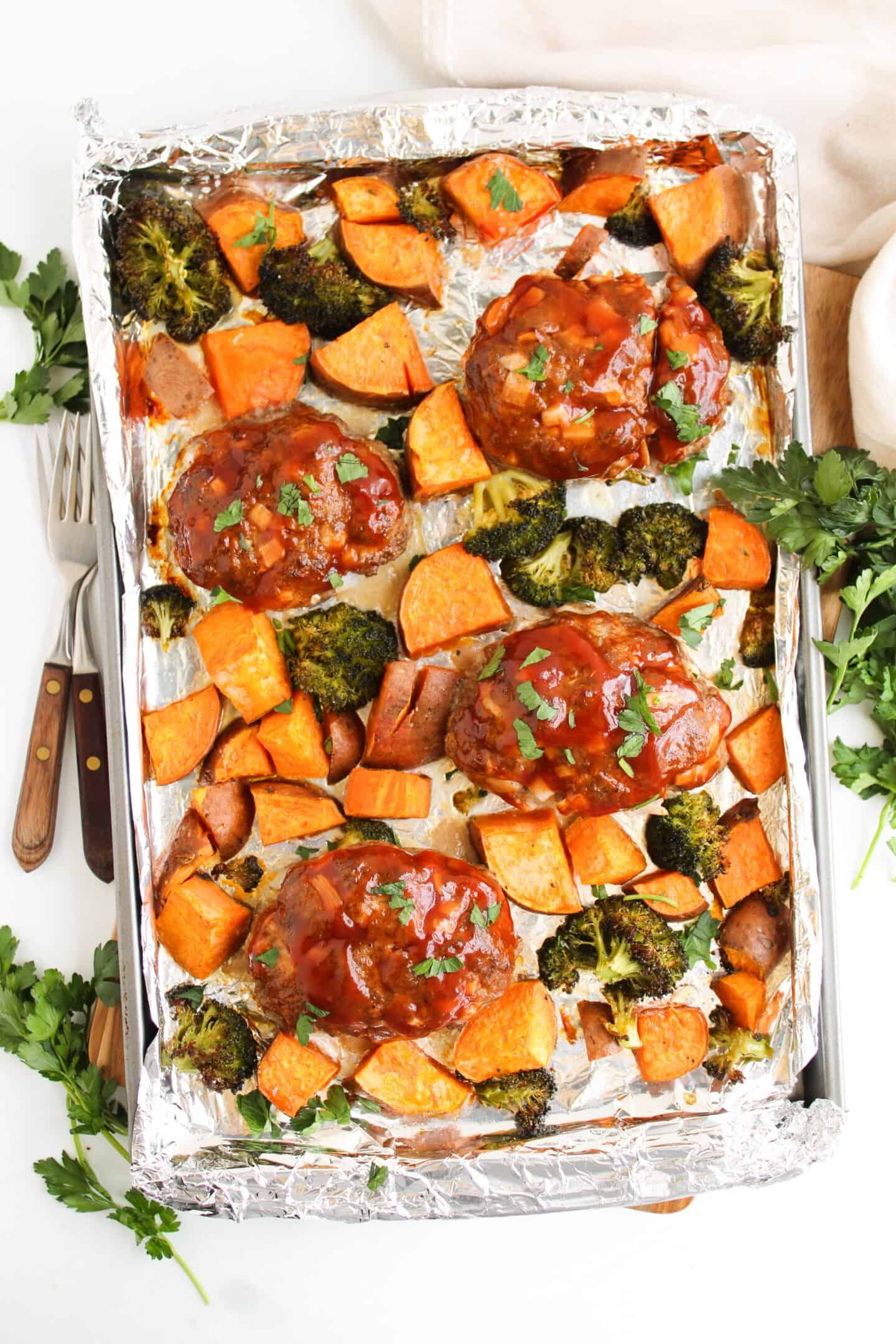 Mini meatloaf portions sit perfectly on a sheet pan, surrounded by roasted veggie goodness—sweet potato chunks and broccoli, all glazed to perfection on a foil-lined surface.