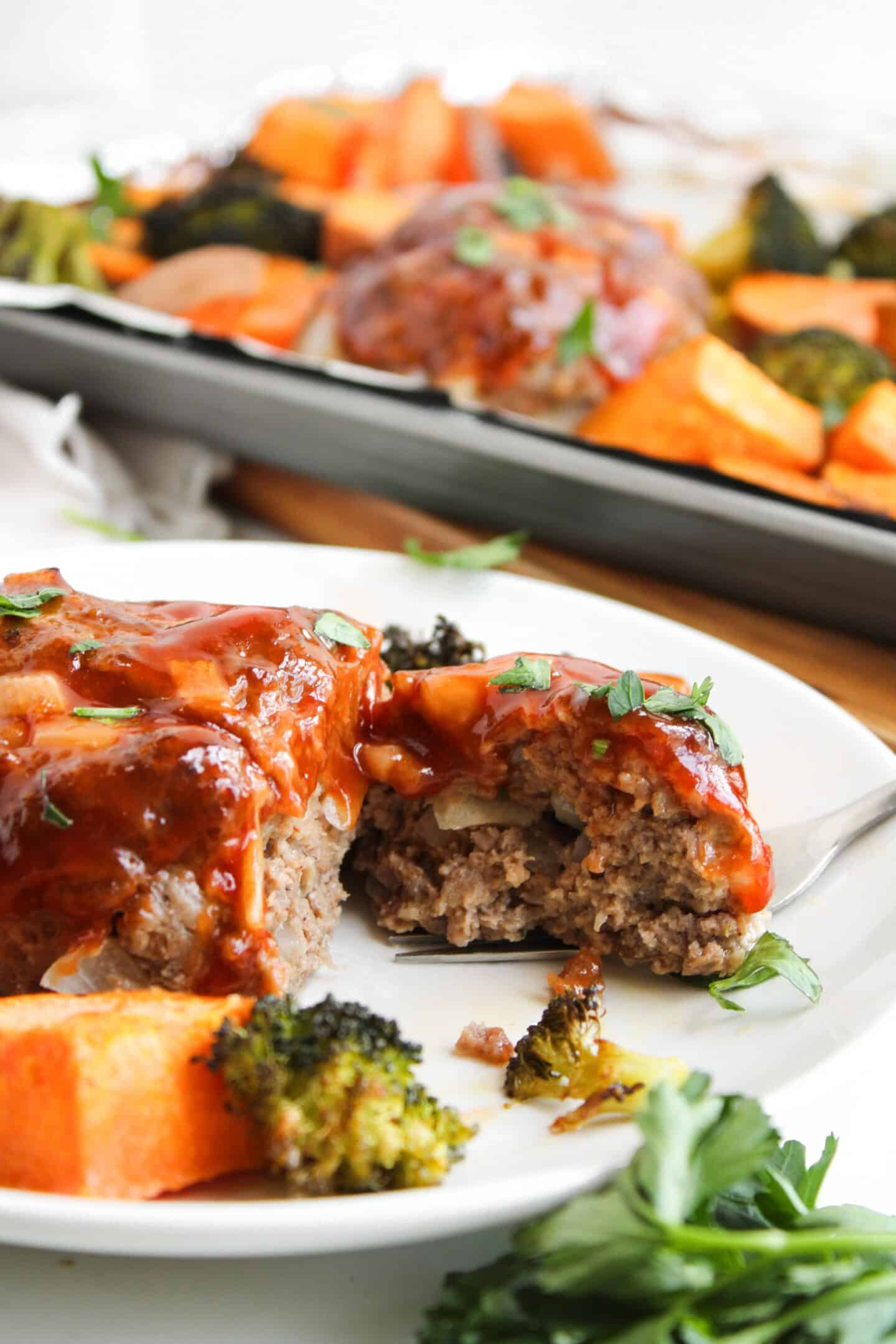 Mini meatloaf topped with sauce sits elegantly on a plate, accompanied by roasted sweet potatoes and vibrant veggies, with a sheet pan resting in the background.