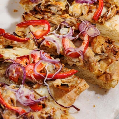 Close-up of BBQ Chicken Flatbread topped with red onion, red bell pepper, shredded chicken, and melted cheese on a white surface.