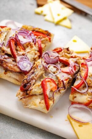 Two slices of BBQ Chicken Flatbread topped with grilled chicken, red bell peppers, red onions, and melted cheese on a cutting board with Swiss cheese slices nearby.