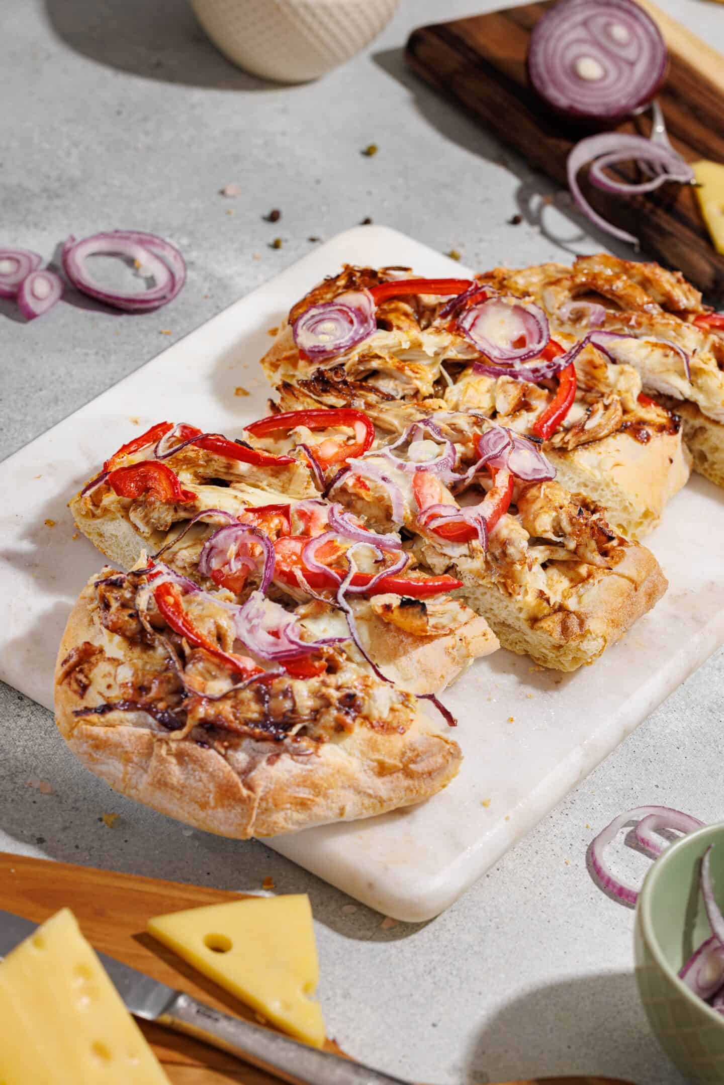 A sliced BBQ chicken flatbread topped with chicken, red bell peppers, and raw red onion sits on a marble board, surrounded by cheese slices and more onion on a gray surface.