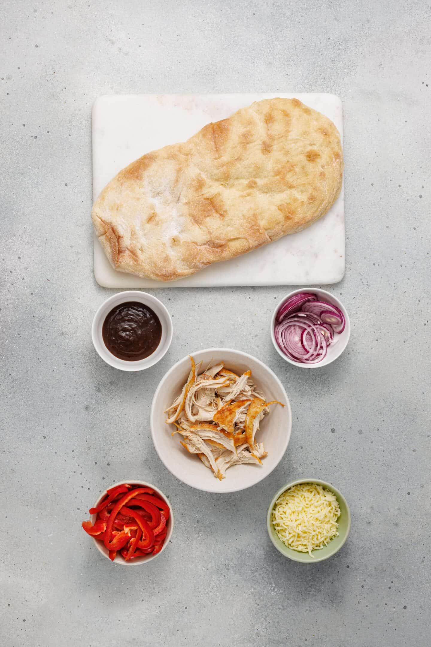 Flatbread on a marble board with bowls of BBQ chicken, tangy BBQ sauce, sliced red onion, sliced red pepper, and grated cheese arranged around it—perfect for building your own BBQ Chicken Flatbread.