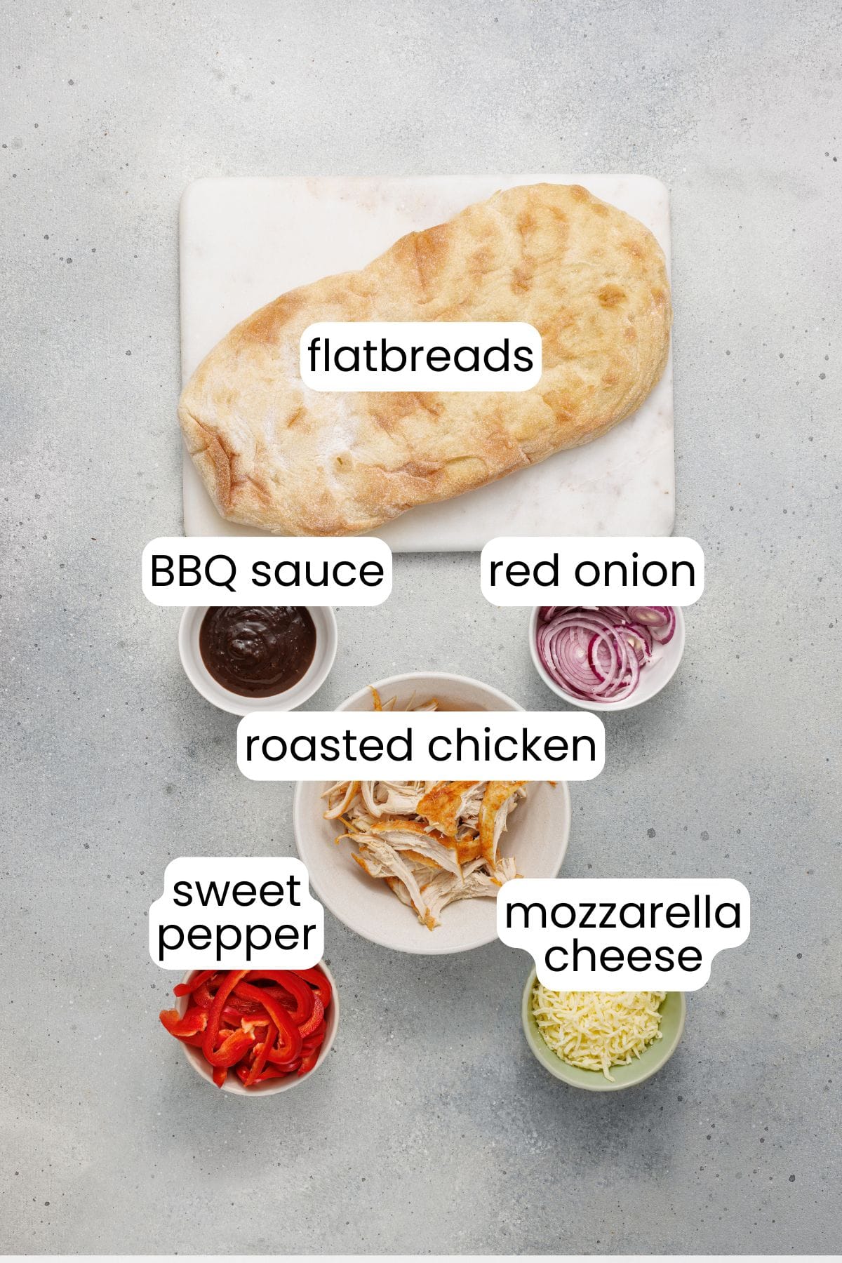 A marble board displays BBQ Chicken Flatbread fixings—bowls of BBQ sauce, red onion, roasted chicken, sweet pepper, and mozzarella cheese—all arranged on a light gray surface.