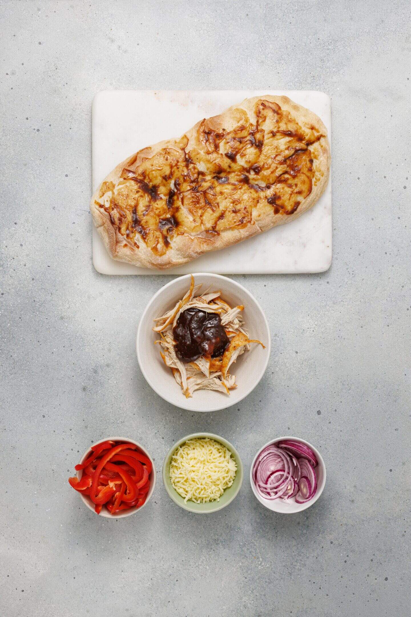 Flatbread with melted cheese and barbecue sauce on a cutting board, with bowls of BBQ chicken, red bell pepper, cheese, and sliced red onion arranged below.
