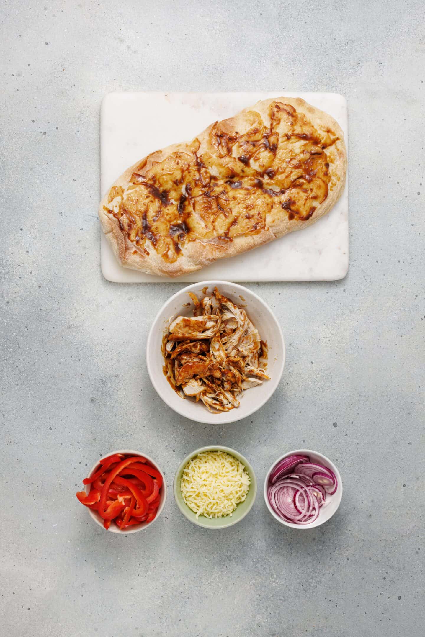 BBQ Chicken Flatbread on a cutting board, surrounded by bowls of shredded chicken, sliced red bell pepper, shredded cheese, and sliced red onion—perfect for assembling your own delicious creation.