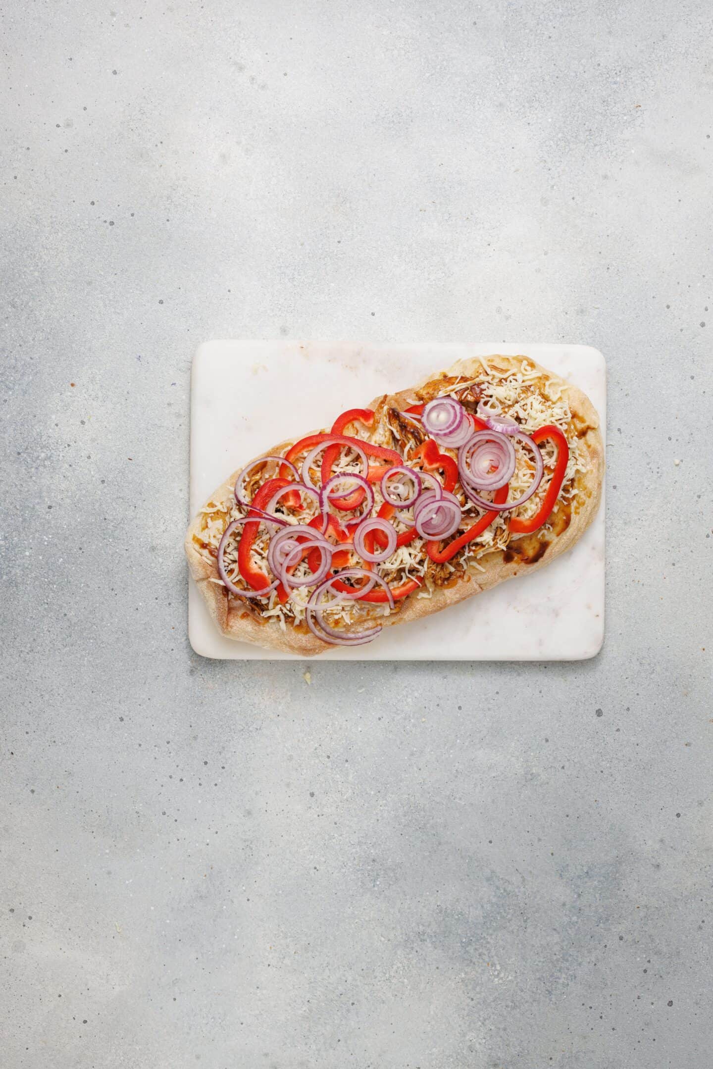 A BBQ chicken flatbread topped with shredded cheese, sliced red bell peppers, and red onions sits on a white rectangular marble board against a light gray background.
