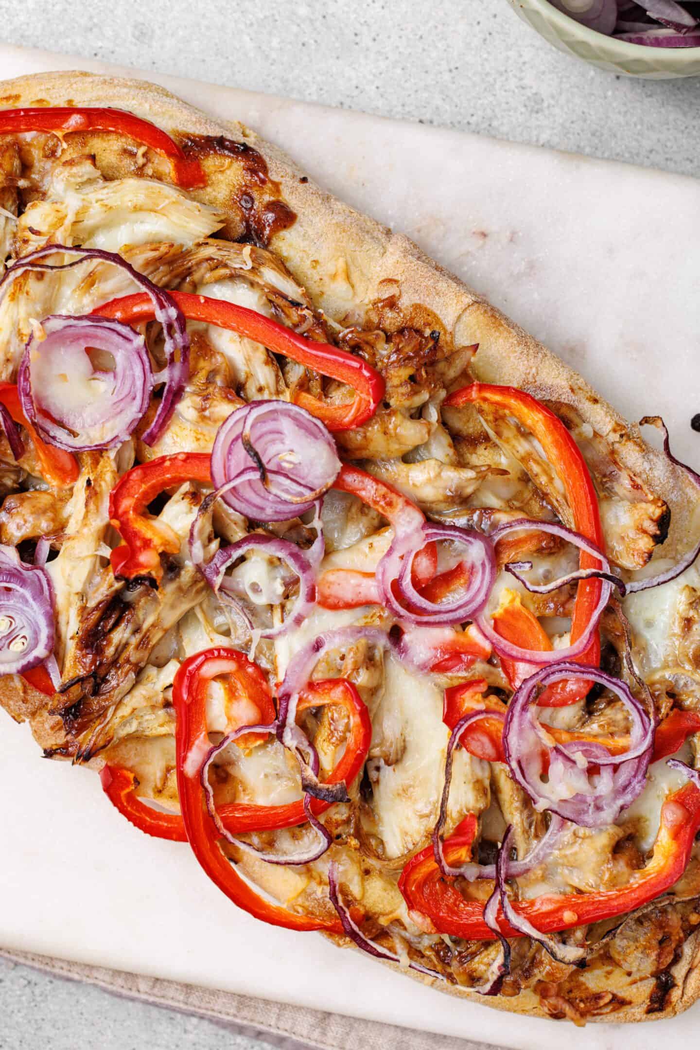 A BBQ Chicken Flatbread topped with shredded chicken, red bell pepper slices, red onion rings, and melted cheese, served on a marble surface.