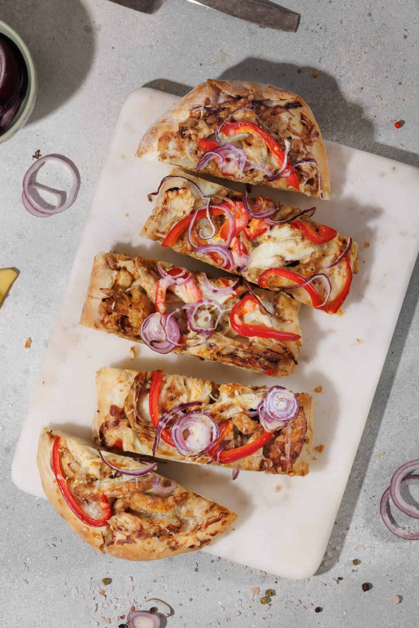 Four rectangular slices of BBQ Chicken Flatbread with red bell pepper, onions, and cheese are arranged on a white cutting board.