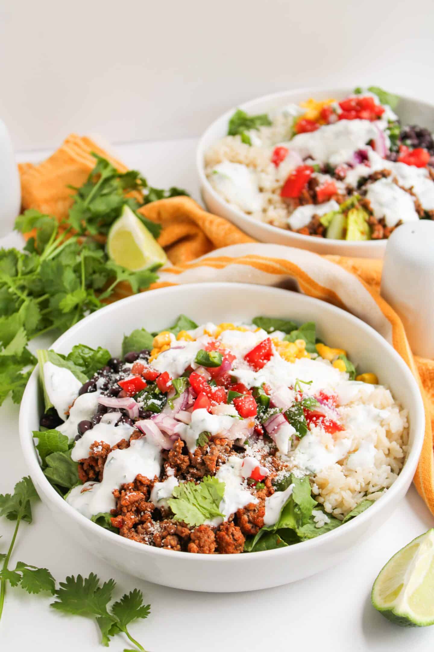 Two Beef Burrito Bowls topped with seasoned ground meat, chopped tomato, corn, red onion, beans, fresh cilantro, and a drizzle of zesty Cilantro Lime Crema, served with lime wedges and herbs on the side.