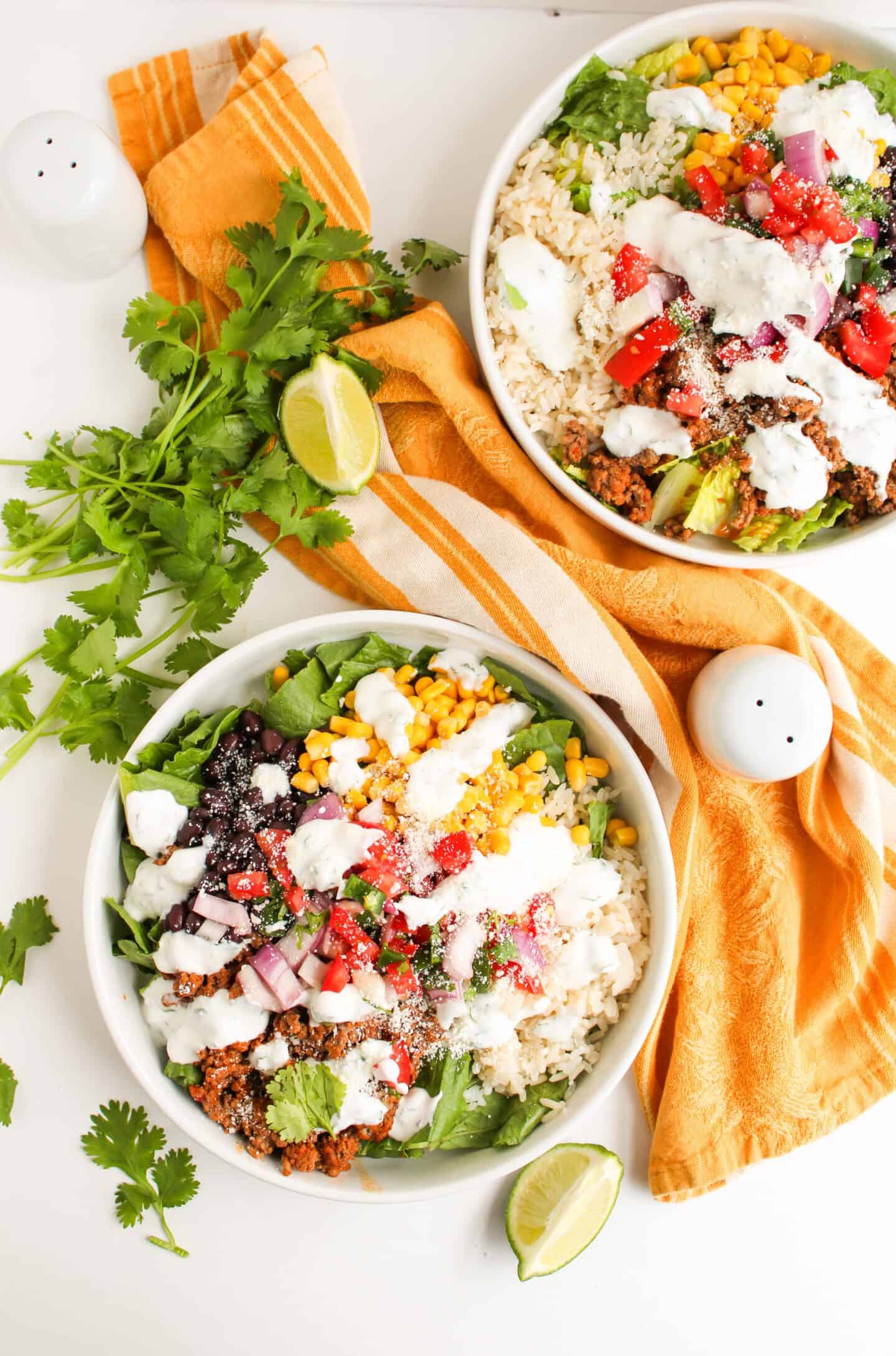 Two bowls of Burrito Bowl salad with rice, lettuce, corn, black beans, onions, tomatoes, and creamy cilantro lime crema dressing, garnished with cilantro and lime on a white table with orange napkins.
