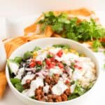 A vibrant Beef Burrito Bowl featuring ground meat, rice, spinach, corn, black beans, diced tomatoes, red onion, cheese, and a drizzle of Cilantro Lime Crema, served with lime wedges and fresh cilantro on the side.