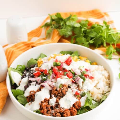A vibrant Beef Burrito Bowl featuring ground meat, rice, spinach, corn, black beans, diced tomatoes, red onion, cheese, and a drizzle of Cilantro Lime Crema, served with lime wedges and fresh cilantro on the side.