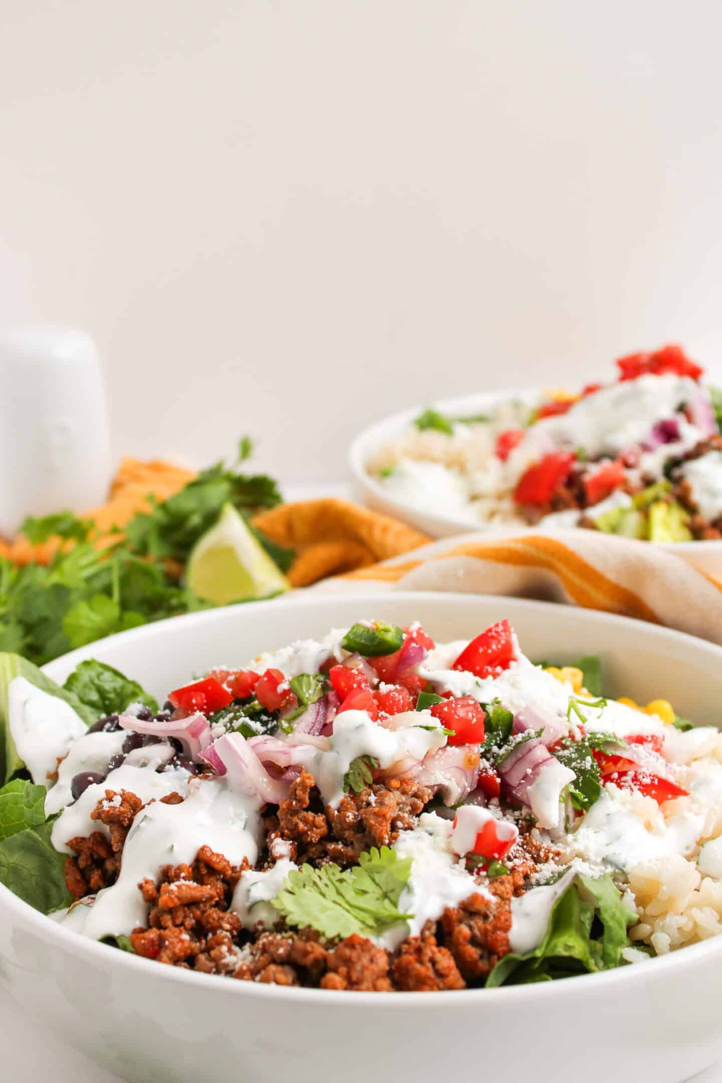 A close-up of a Burrito Bowl with lettuce, seasoned ground meat, diced tomatoes, onions, cilantro, and a drizzle of Cilantro Lime Crema, with another similar bowl in the background.