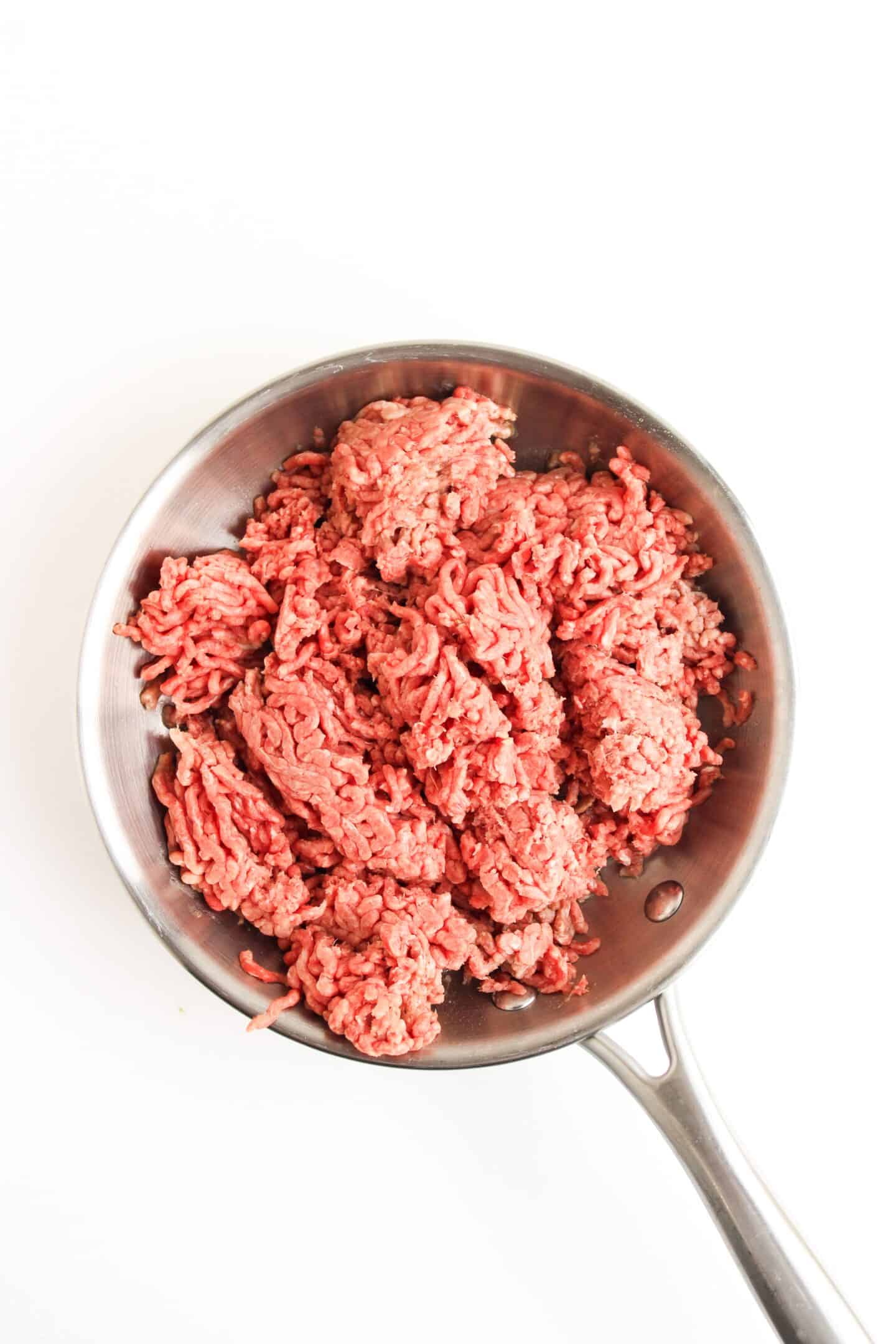 Raw ground beef in a metal skillet on a white background, ready to be transformed into a flavorful Beef Burrito Bowl or paired with zesty cilantro lime crema.
