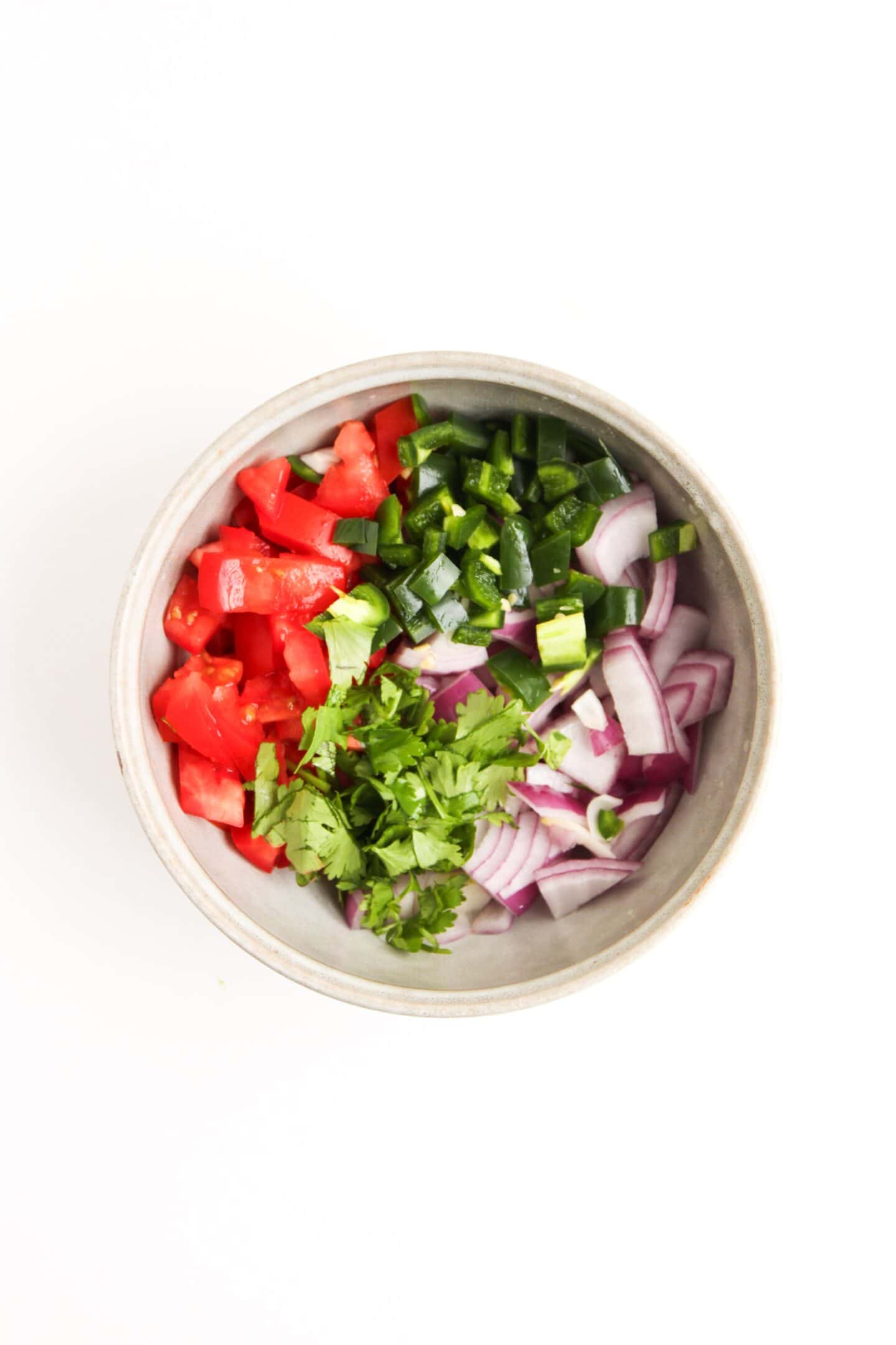 A bowl containing chopped tomatoes, jalapeños, red onions, and cilantro, arranged in separate sections on a white background—perfect fresh toppings for a Beef Burrito Bowl or to pair with Cilantro Lime Crema.