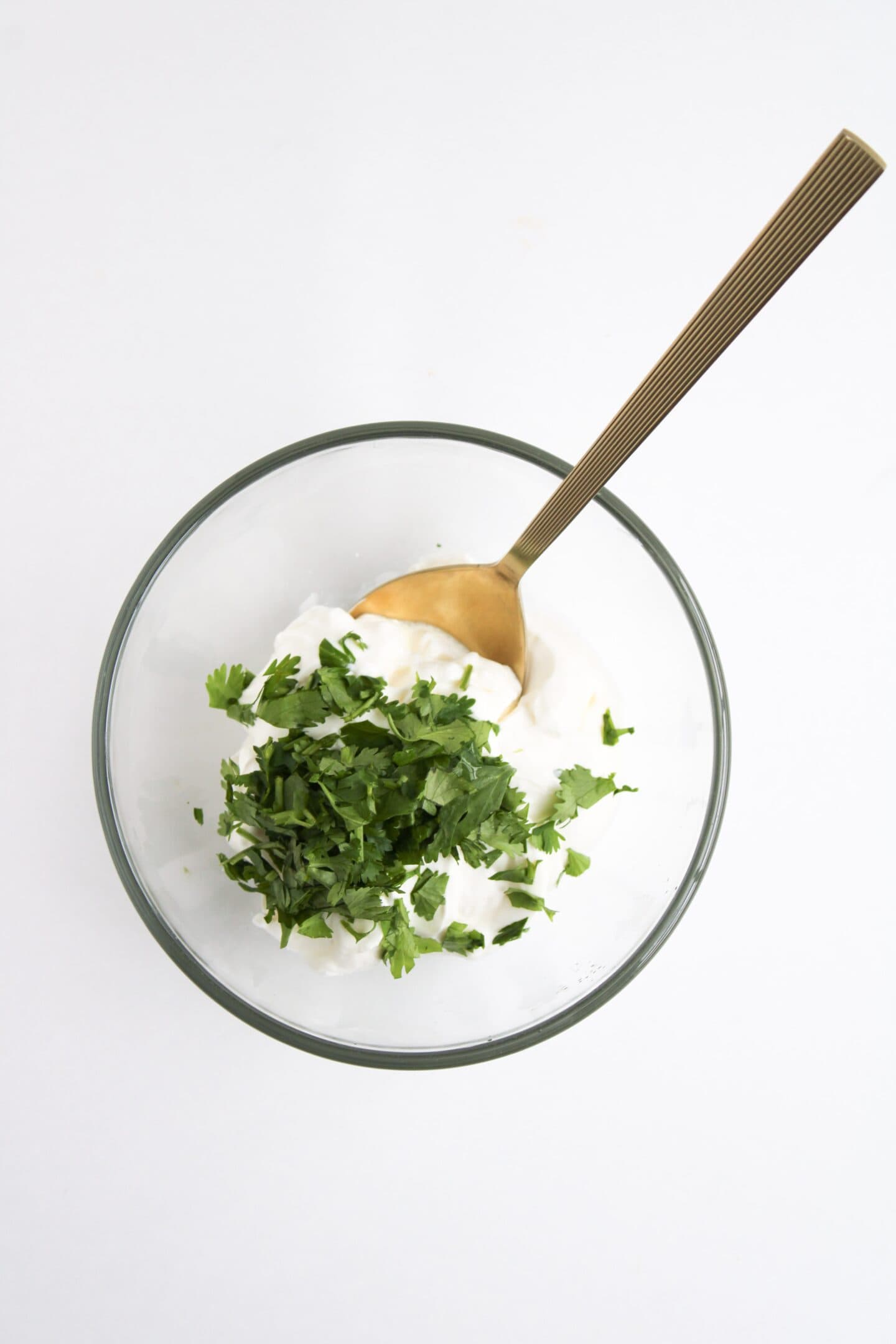A glass bowl with Cilantro Lime Crema and chopped cilantro, with a gold spoon resting inside, perfect for drizzling over your favorite Burrito Bowl on a white background.