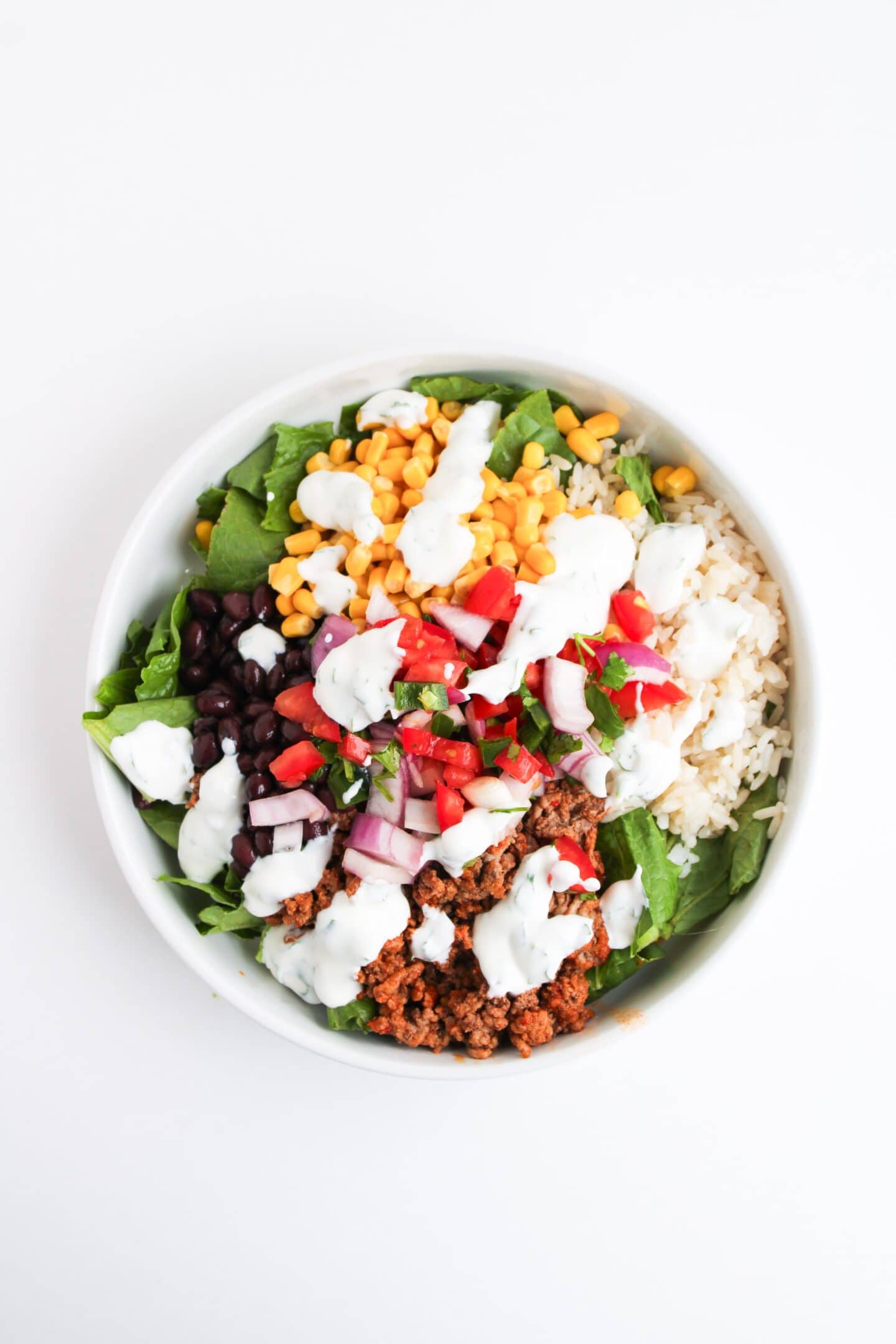 A Beef Burrito Bowl with rice, seasoned ground meat, black beans, corn, spinach, diced tomatoes, red onions, and a drizzle of tangy Cilantro Lime Crema on top.