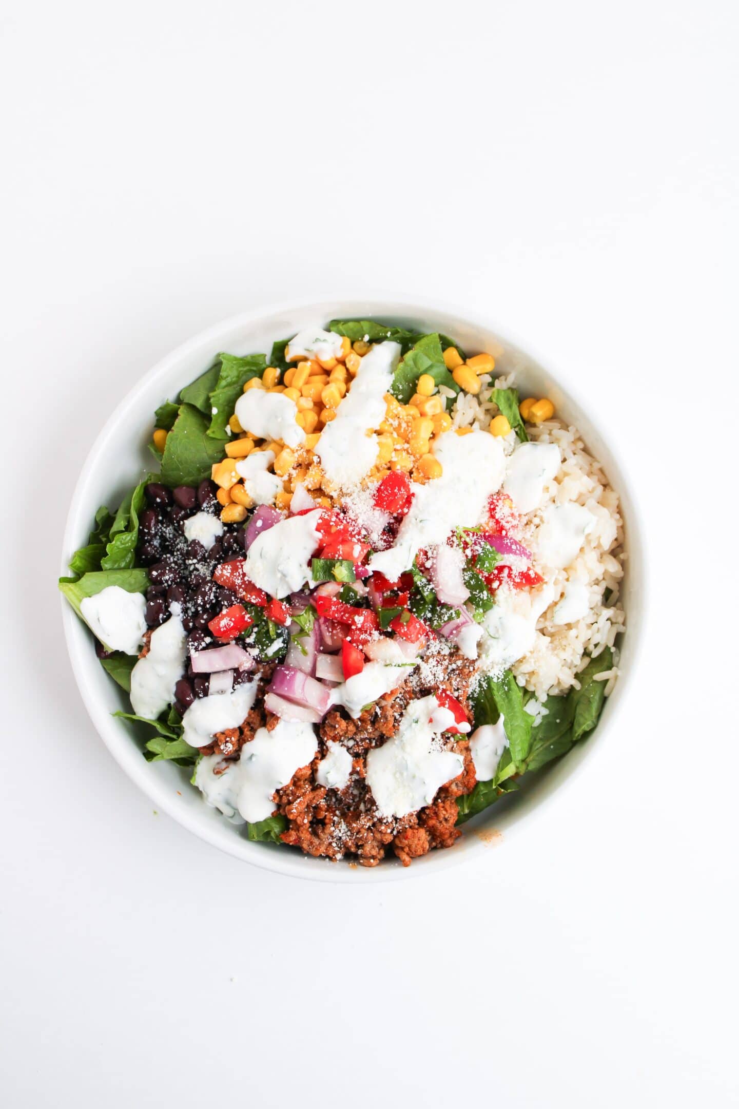 A Beef Burrito Bowl with ground beef, rice, black beans, corn, spinach, diced tomatoes, red onions, shredded cheese, and drizzled with cilantro lime crema in a white bowl.
