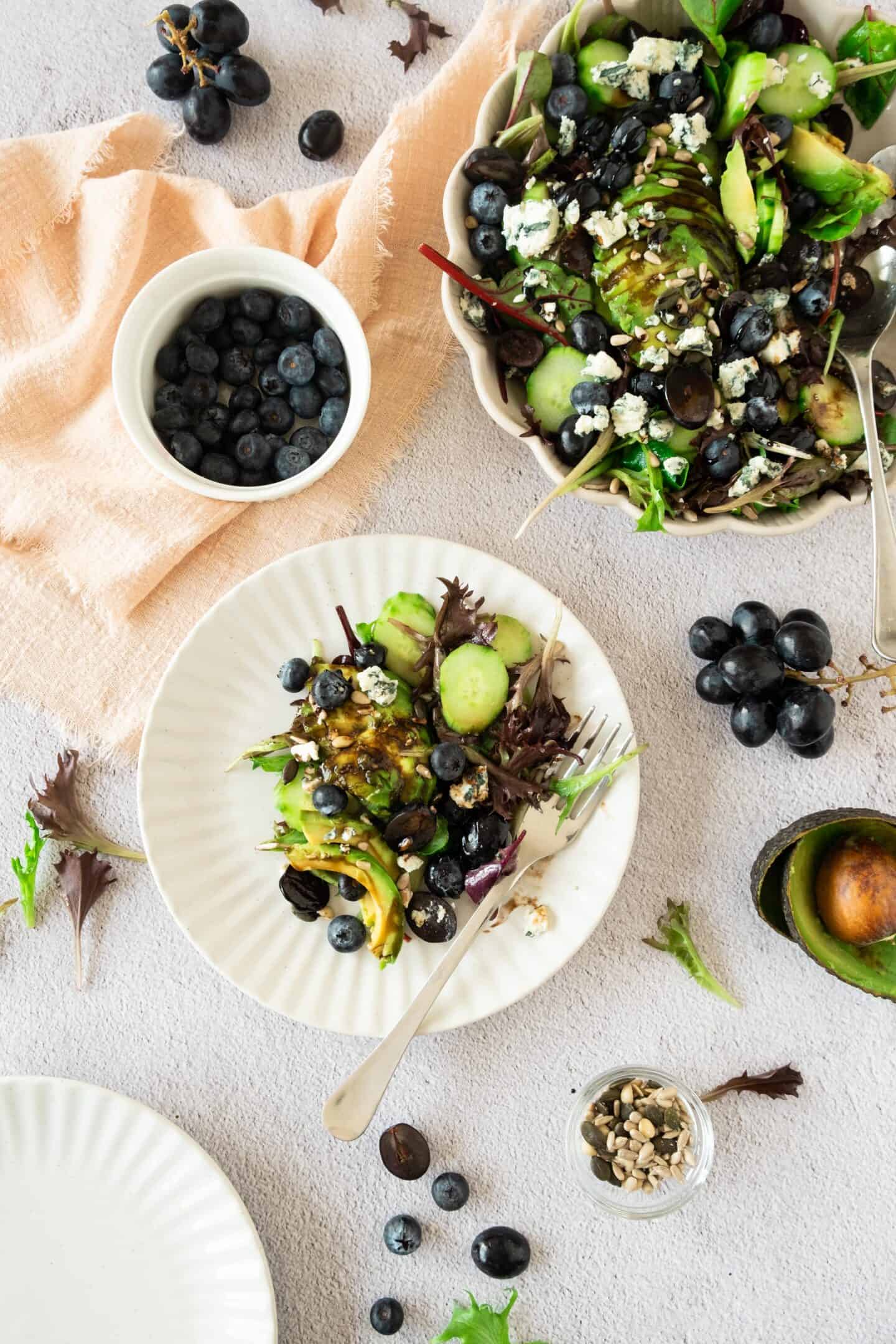 A vibrant plate of mixed green salad with creamy avocado, fresh blueberry and grape, seeds, and cheese, accompanied by bowls of blueberries and salad on a light textured surface.