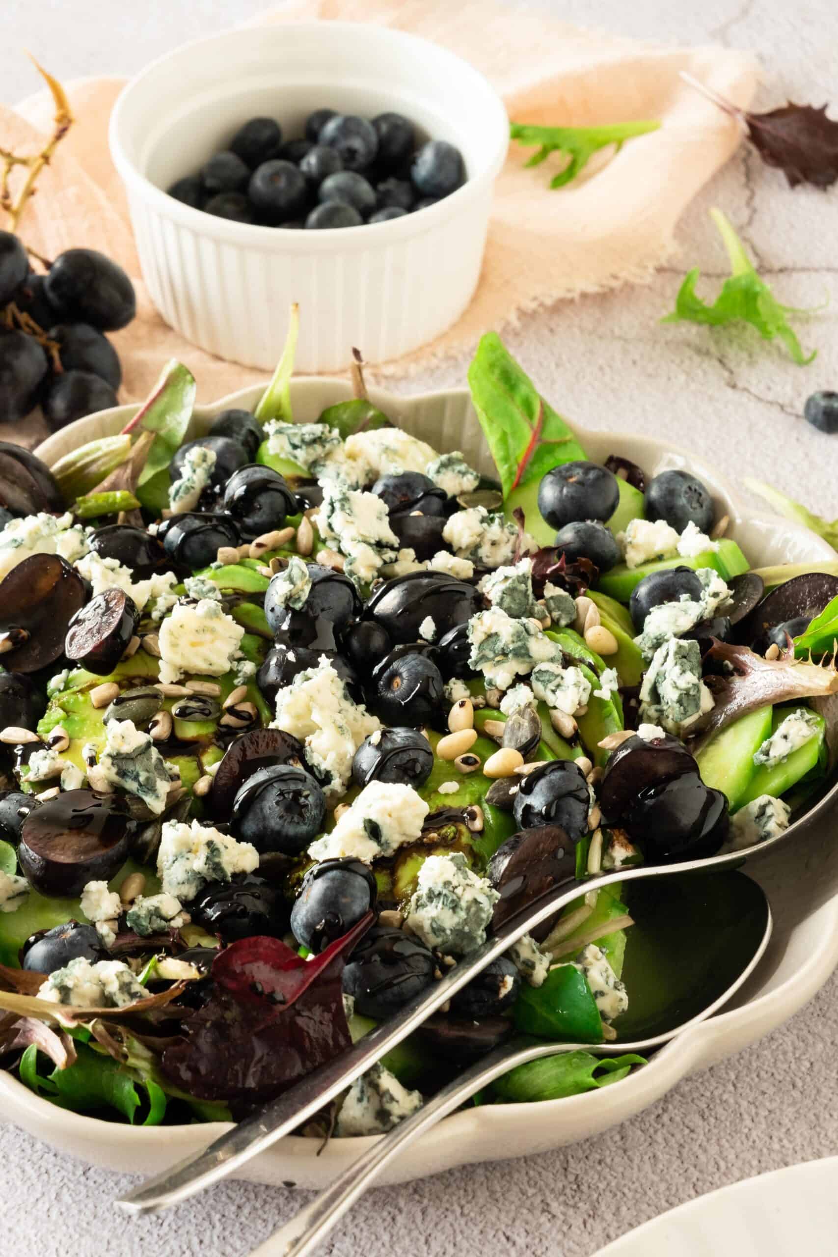 A bowl of mixed green salad topped with blue cheese, grape halves, blueberries, and sunflower seeds, with a serving spoon beside it. A small bowl of fresh blueberries sits in the background.