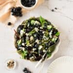 A bowl of salad with leafy greens, blueberries, black grapes, and crumbled cheese sits beside a small bowl of blueberries and seeds, with an empty plate and utensils nearby.