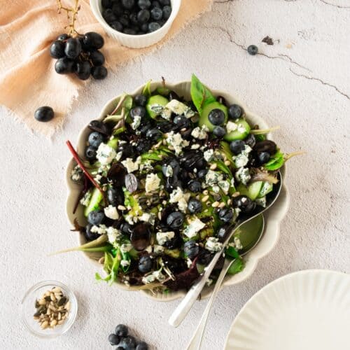 A bowl of salad with leafy greens, blueberries, black grapes, and crumbled cheese sits beside a small bowl of blueberries and seeds, with an empty plate and utensils nearby.